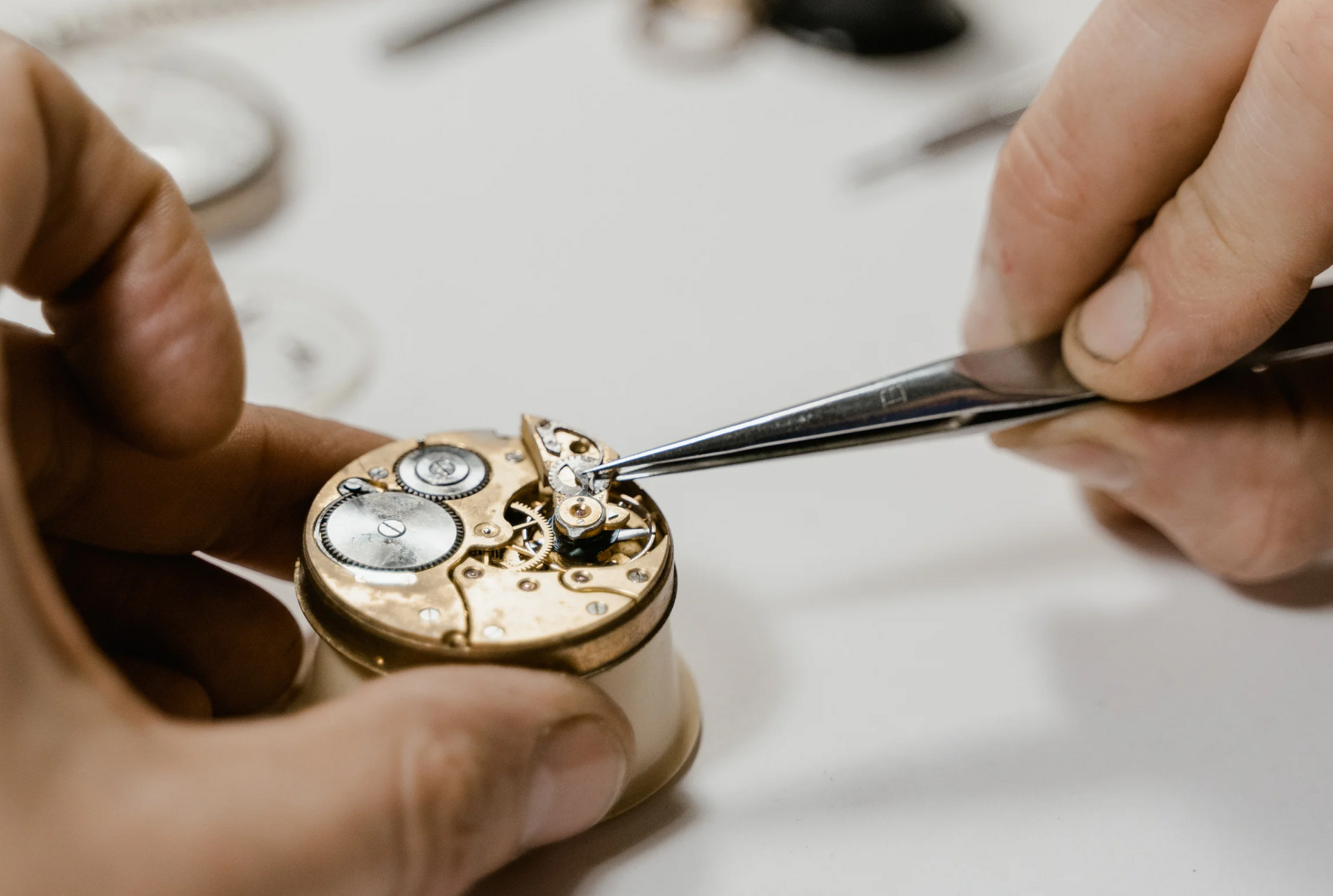 Hands repairing a small watch mechanism with tweezers on a white surface.