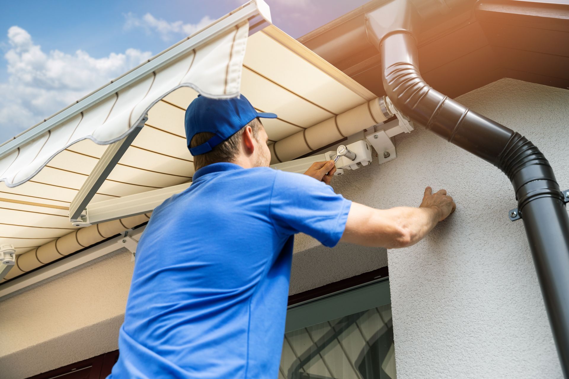 Worker in blue shirt cleaning an exterior wall under a roof eave with a brush and hose