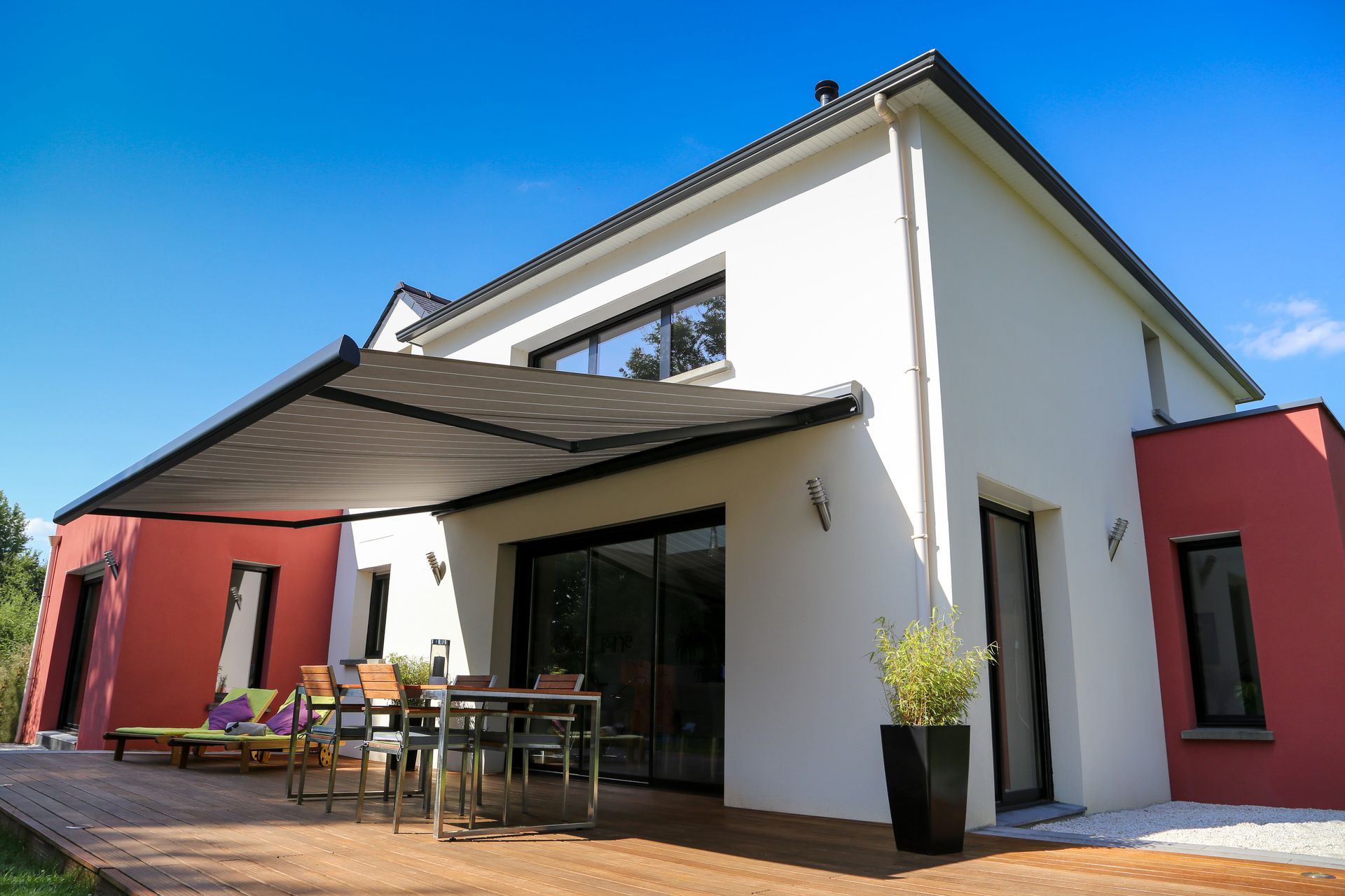 Modern white house with red accents, patio furniture, and a large shaded terrace under a blue sky
