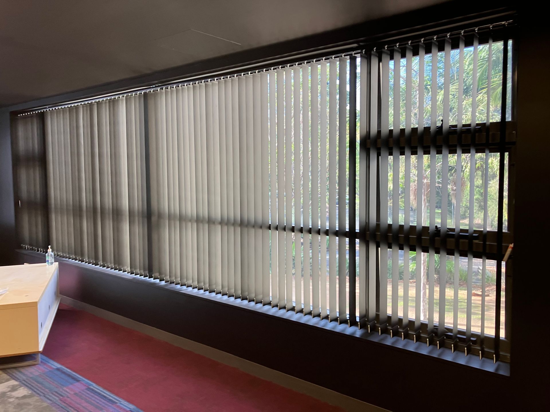 Empty room with large windows, vertical blinds, and sunlight casting shadows on the wall and carpet.