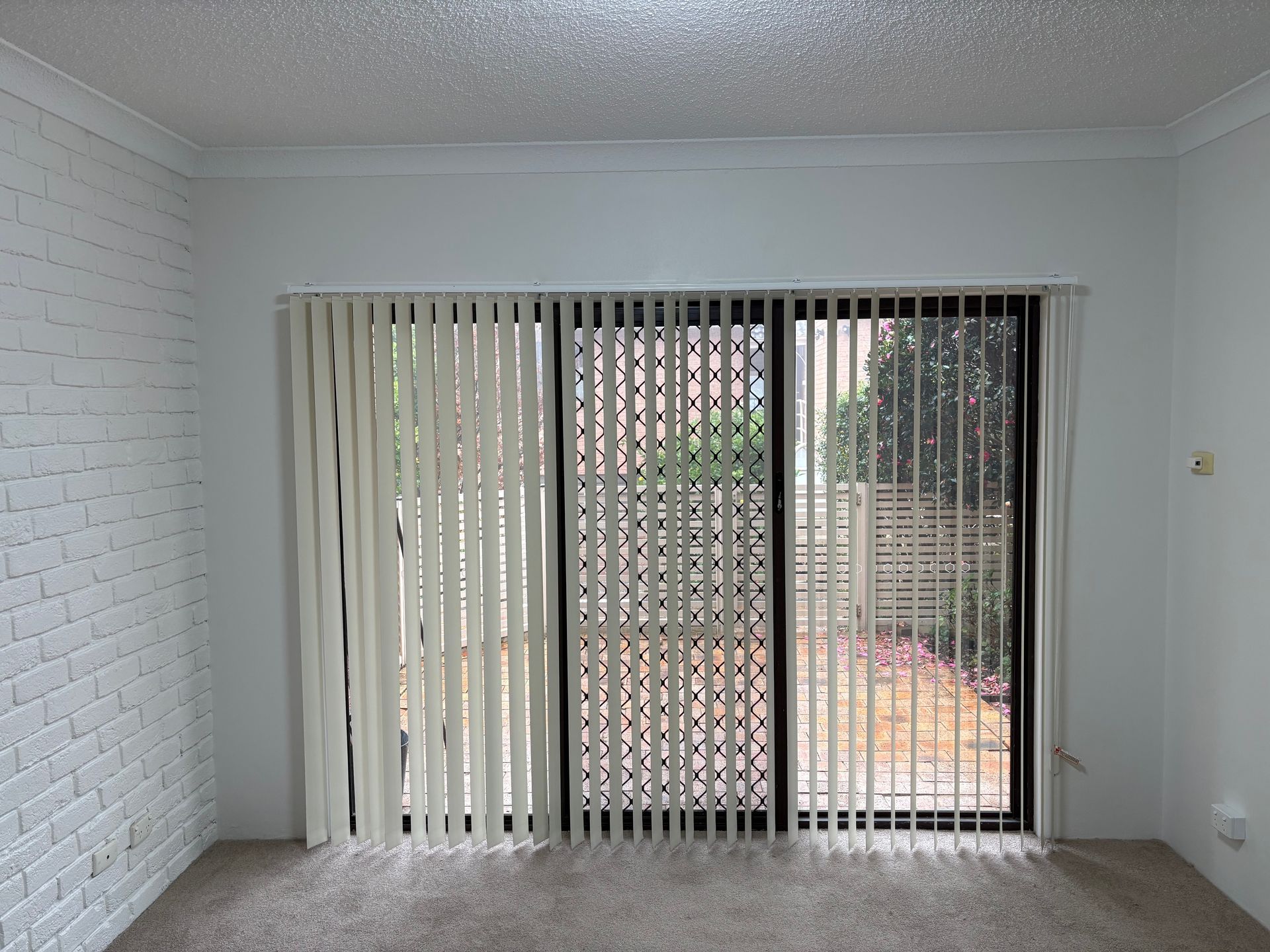 Empty living room with white brick wall, vertical blinds, and sliding glass door overlooking greenery