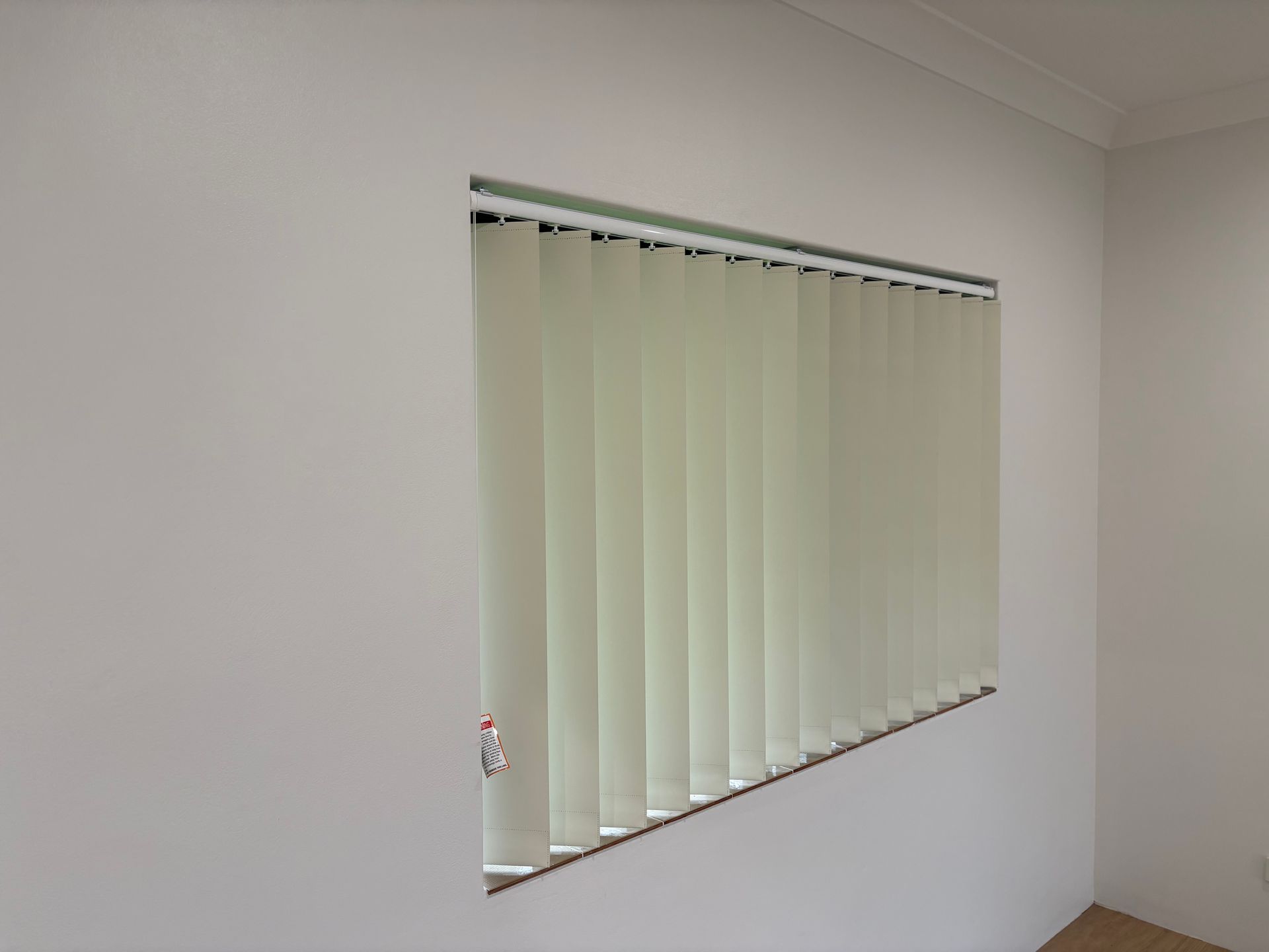 Window with closed white vertical blinds in a plain room.