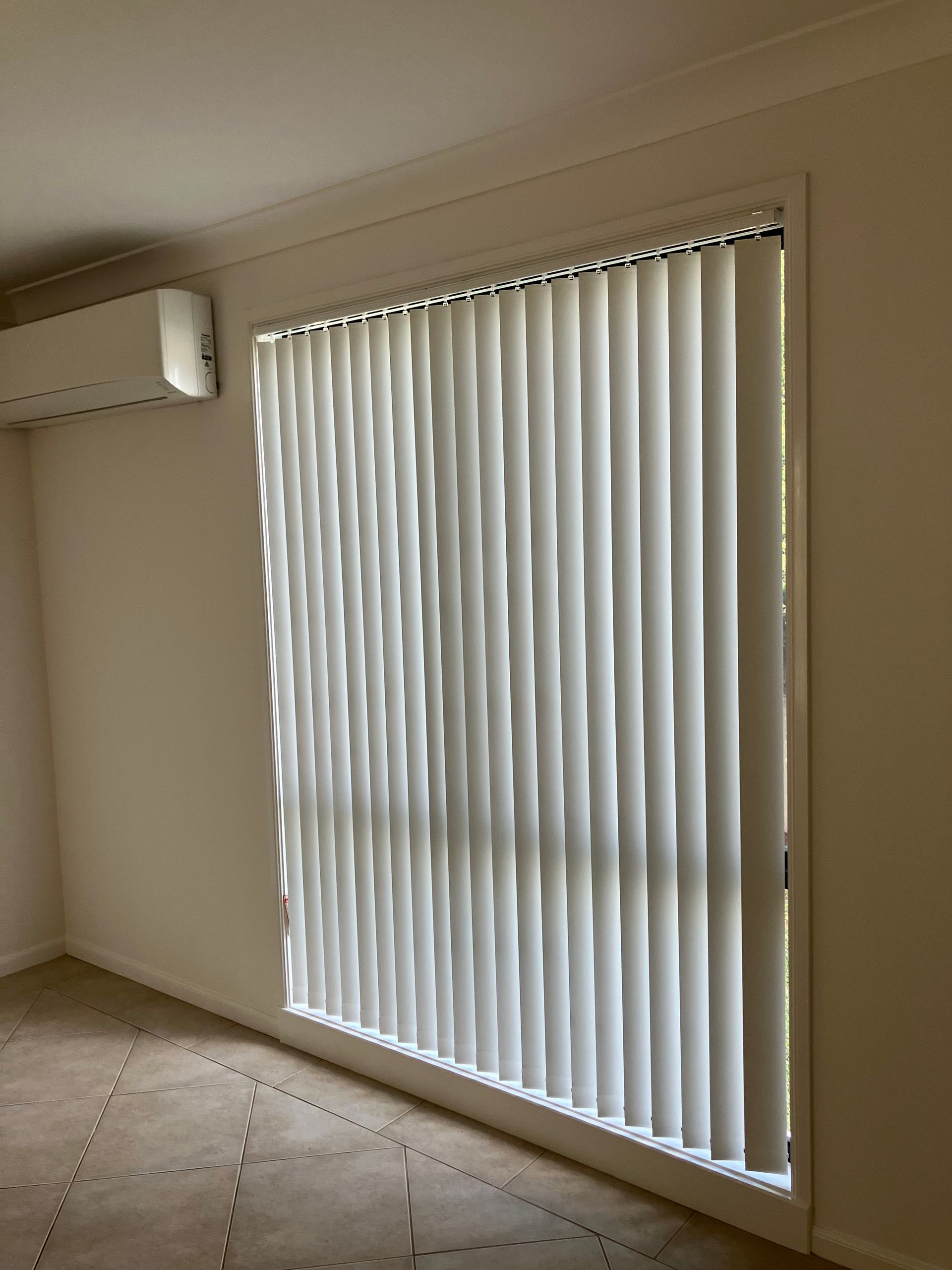 Empty room with beige walls, wall-mounted AC, and vertical blinds covering a large window.