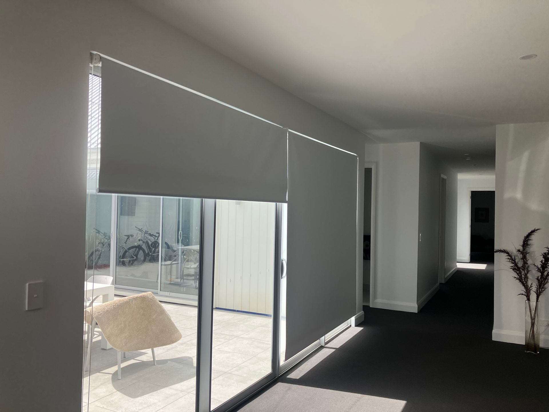 Modern living room with gray blinds over sliding glass doors, dark carpet, and a hallway to the right