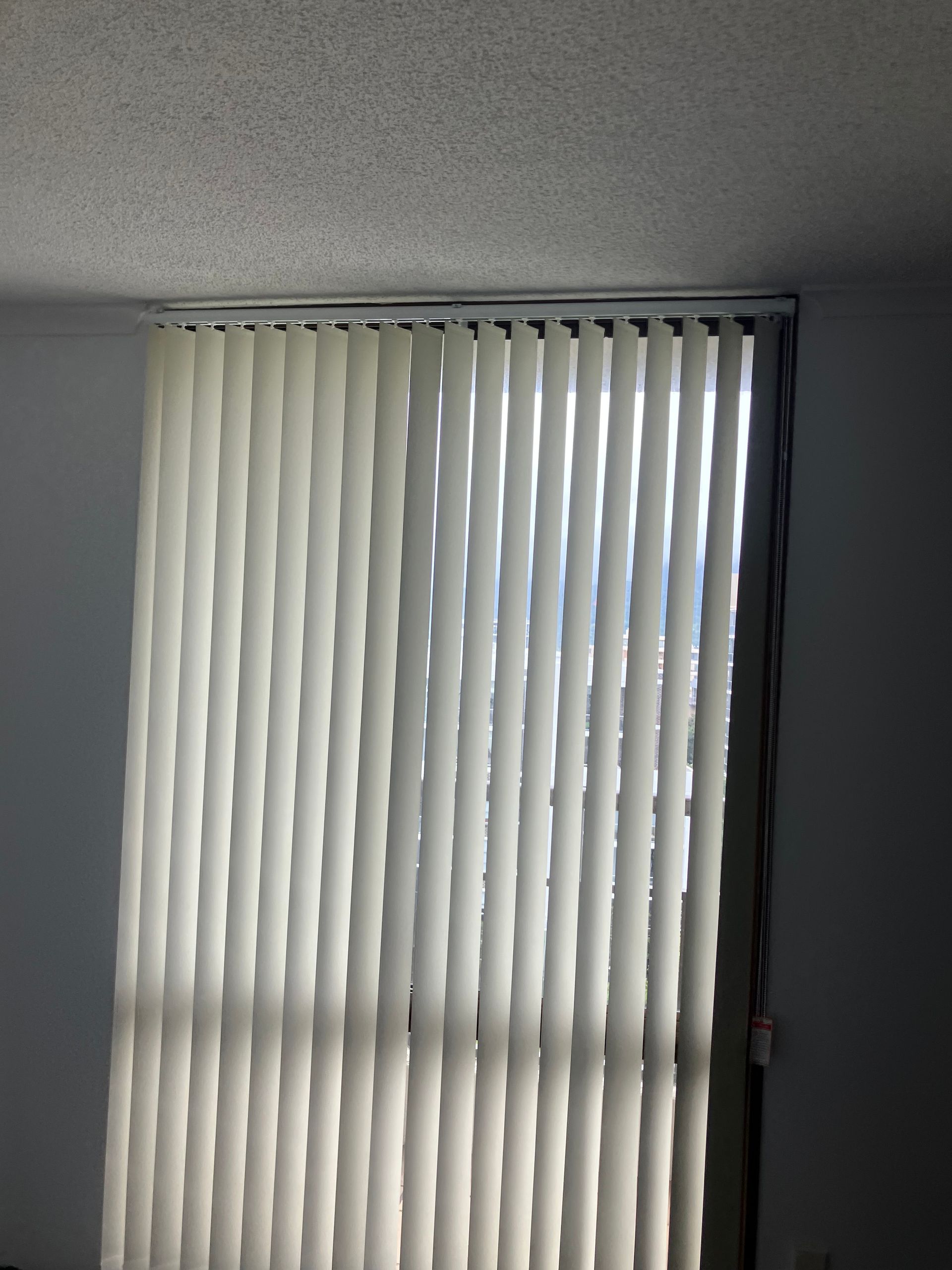Closed white vertical blinds covering a window in a room, with light peeking through gaps.