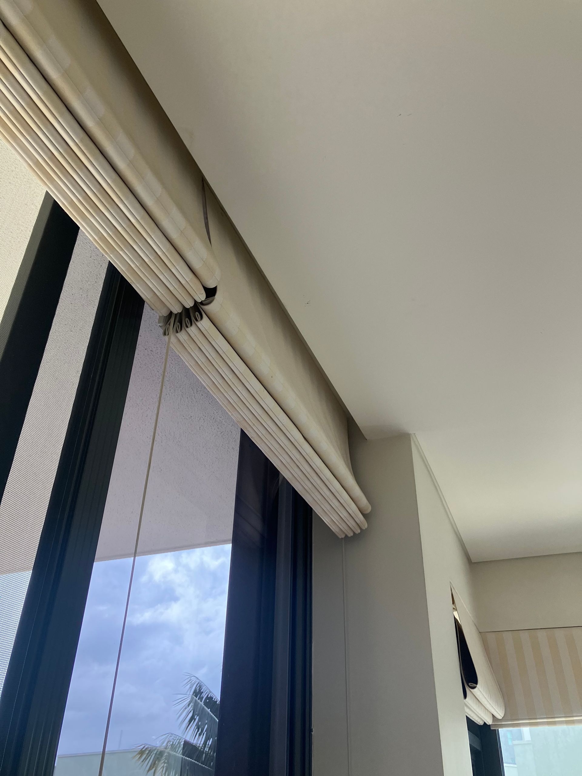 Interior window with beige roman blinds and sheer curtains, showing a cloudy sky outside
