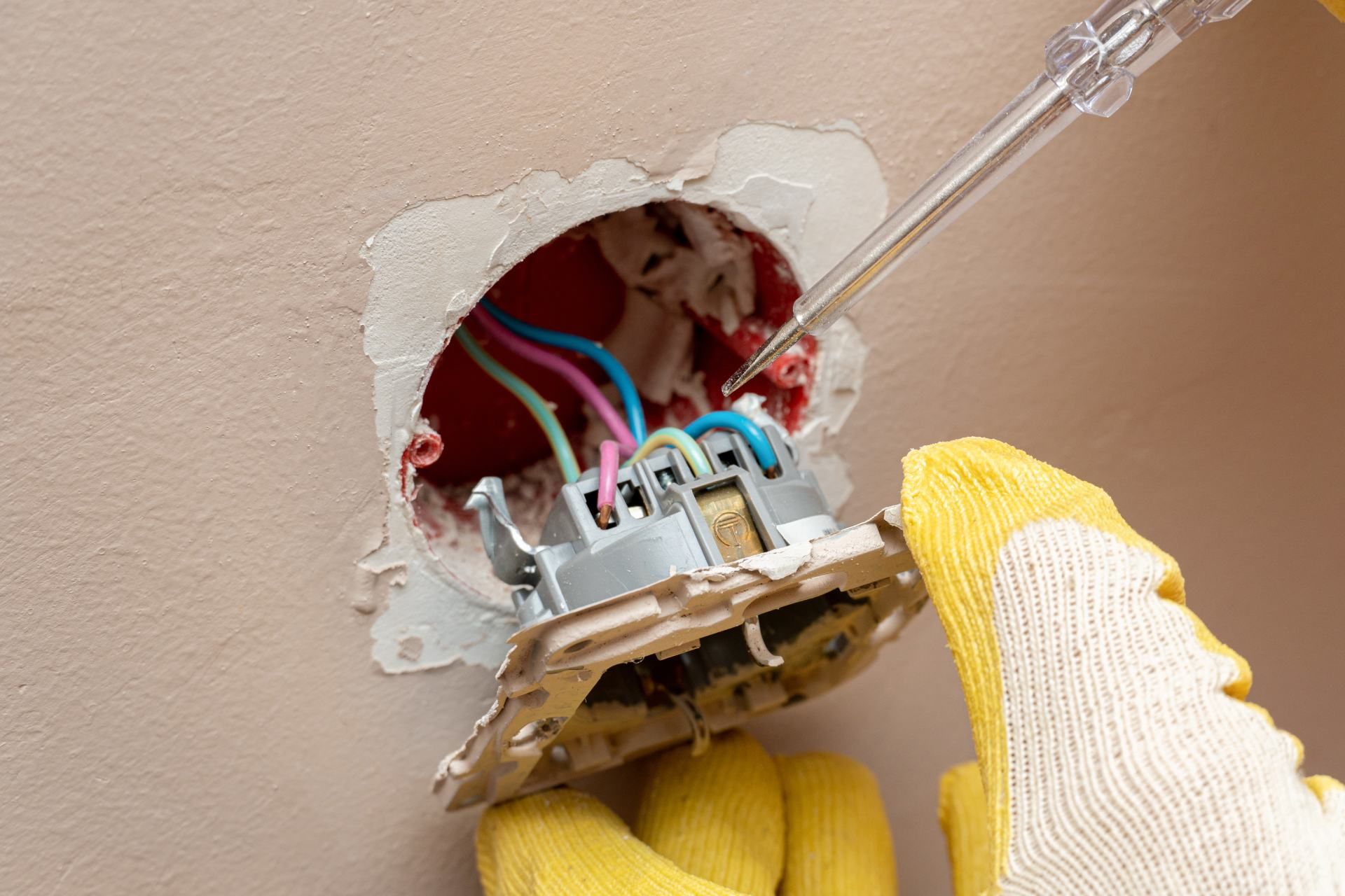 Electrician testing wires in an open wall outlet, wearing yellow gloves, using a voltage tester.