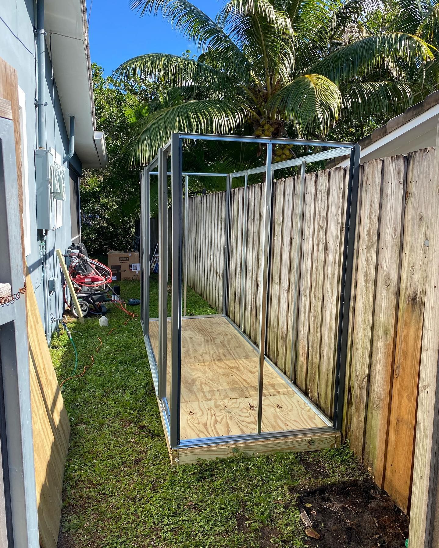 Frame of a shed under construction between a building and a wooden fence on grass.