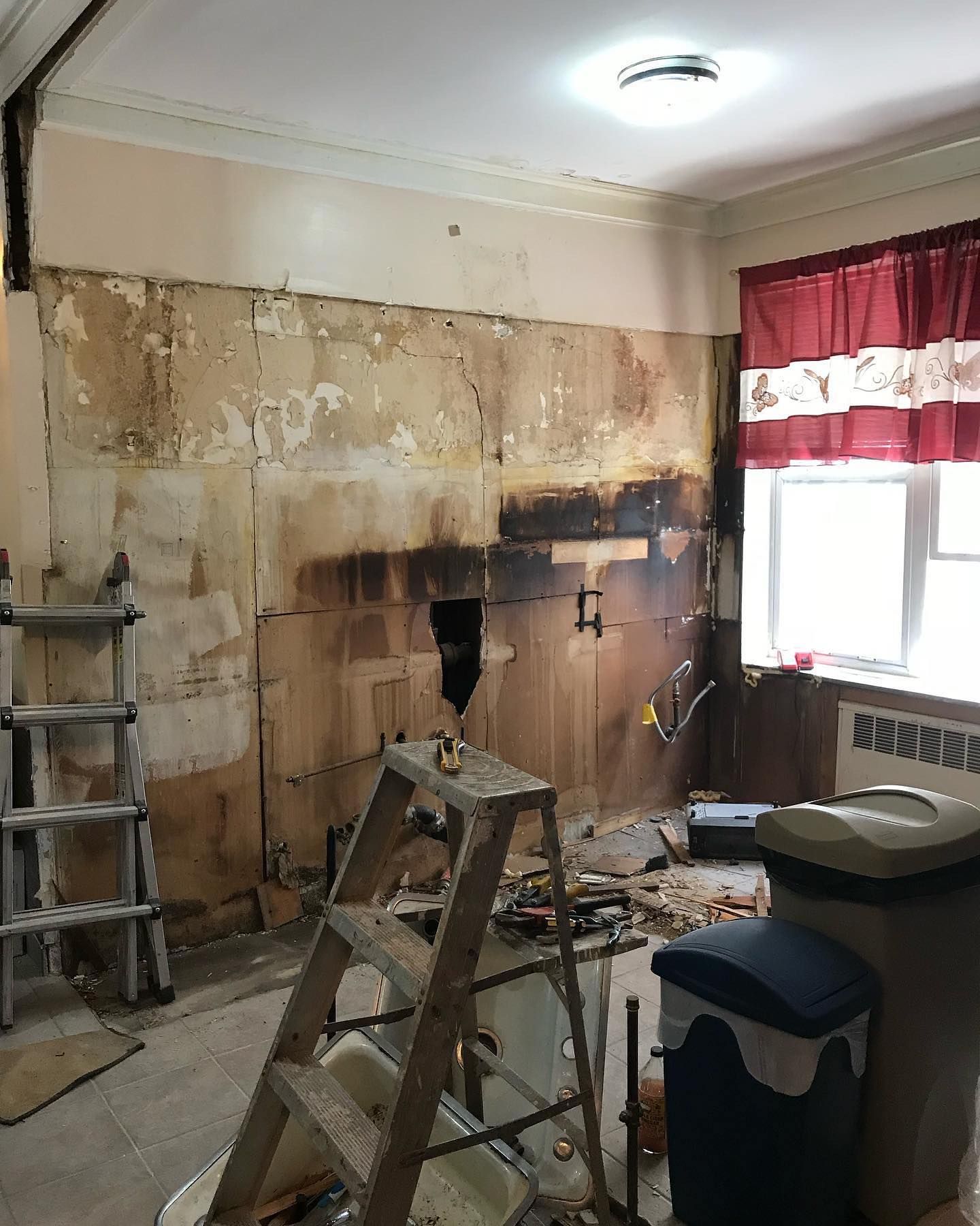 Kitchen under renovation, with exposed wall, tools, ladder, trash bins, and window with curtains.