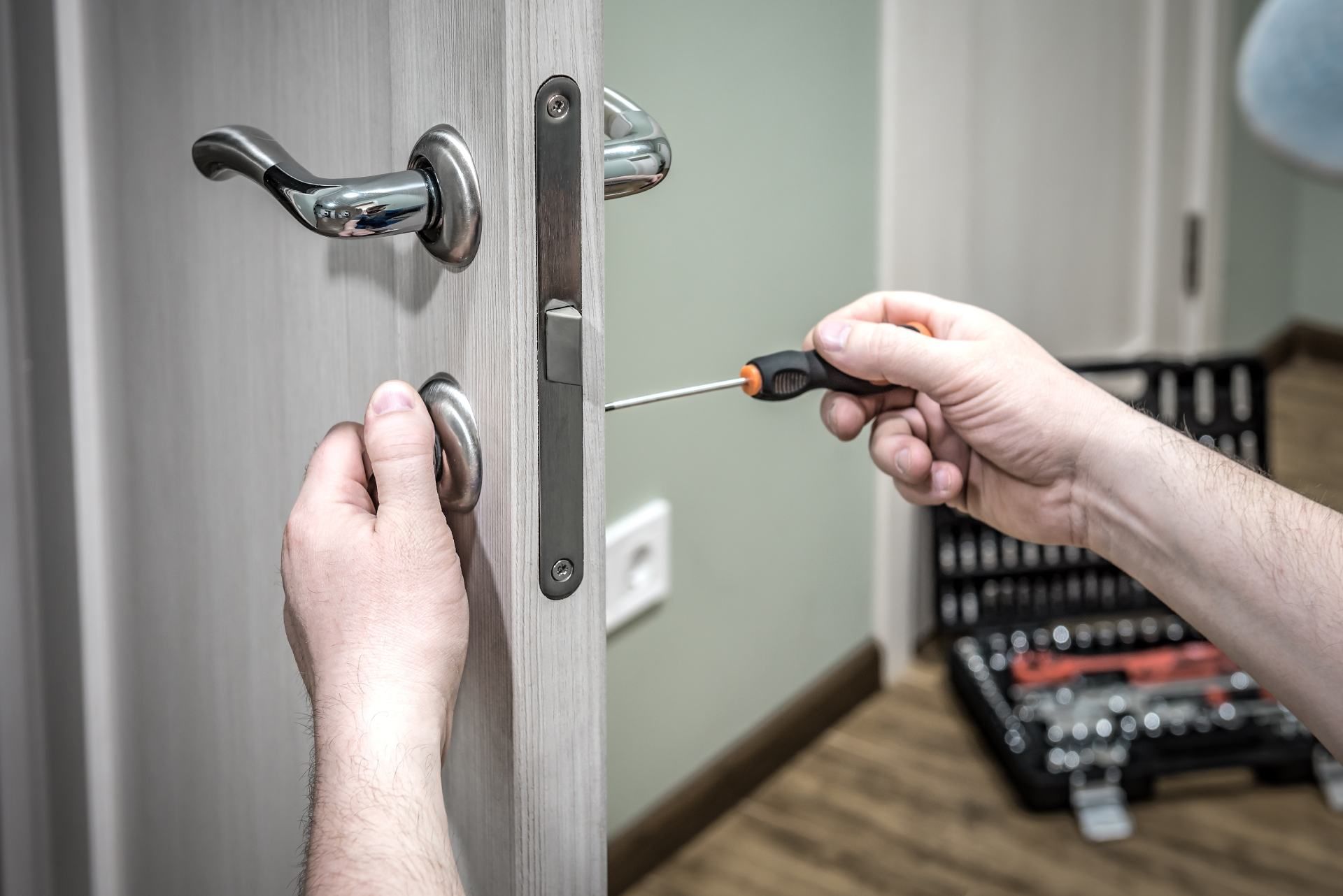 Person repairing a door lock with a screwdriver, a tool set nearby.