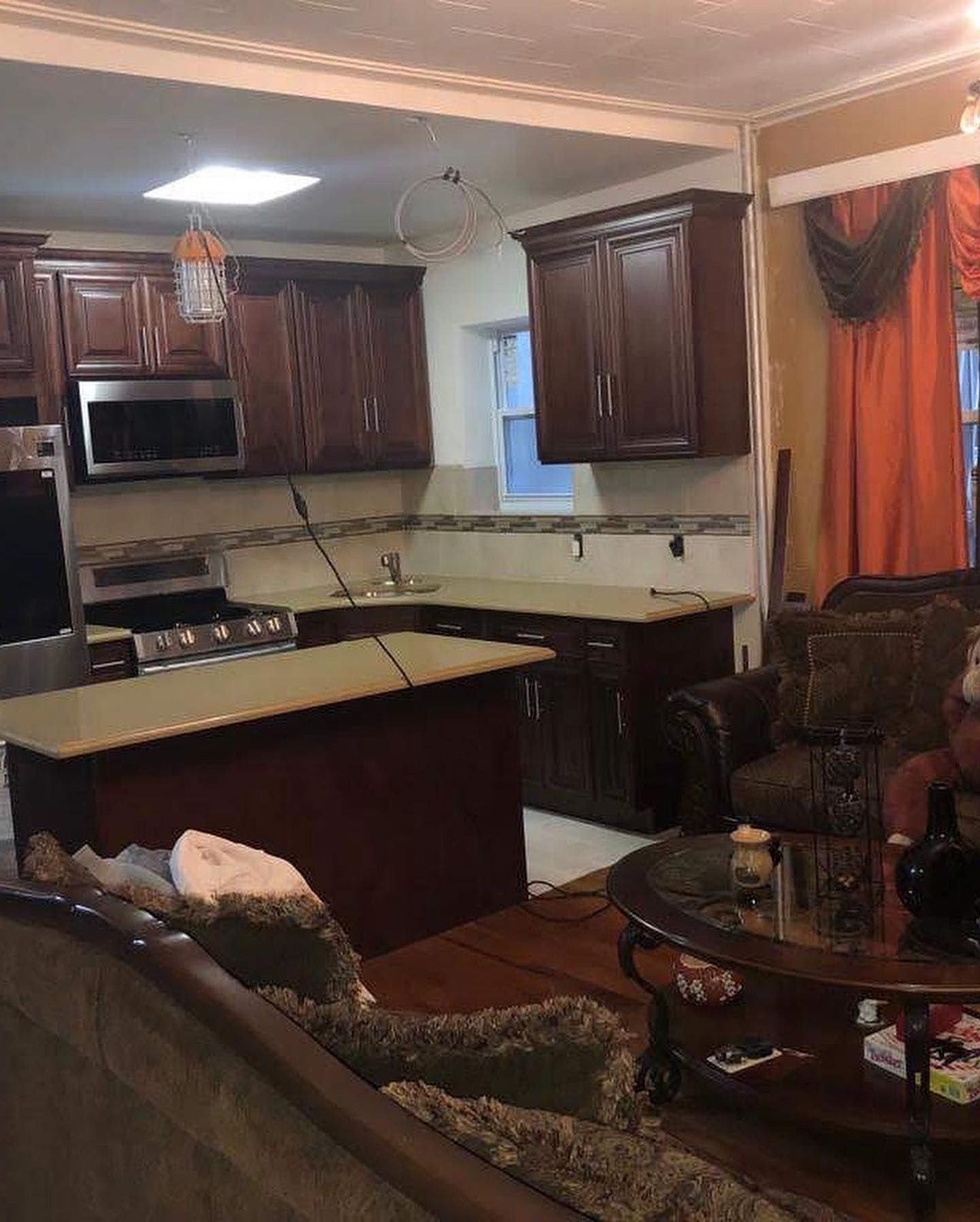Dark wood kitchen with island, brown cabinets, tan countertops, stainless steel appliances, and adjacent living room.