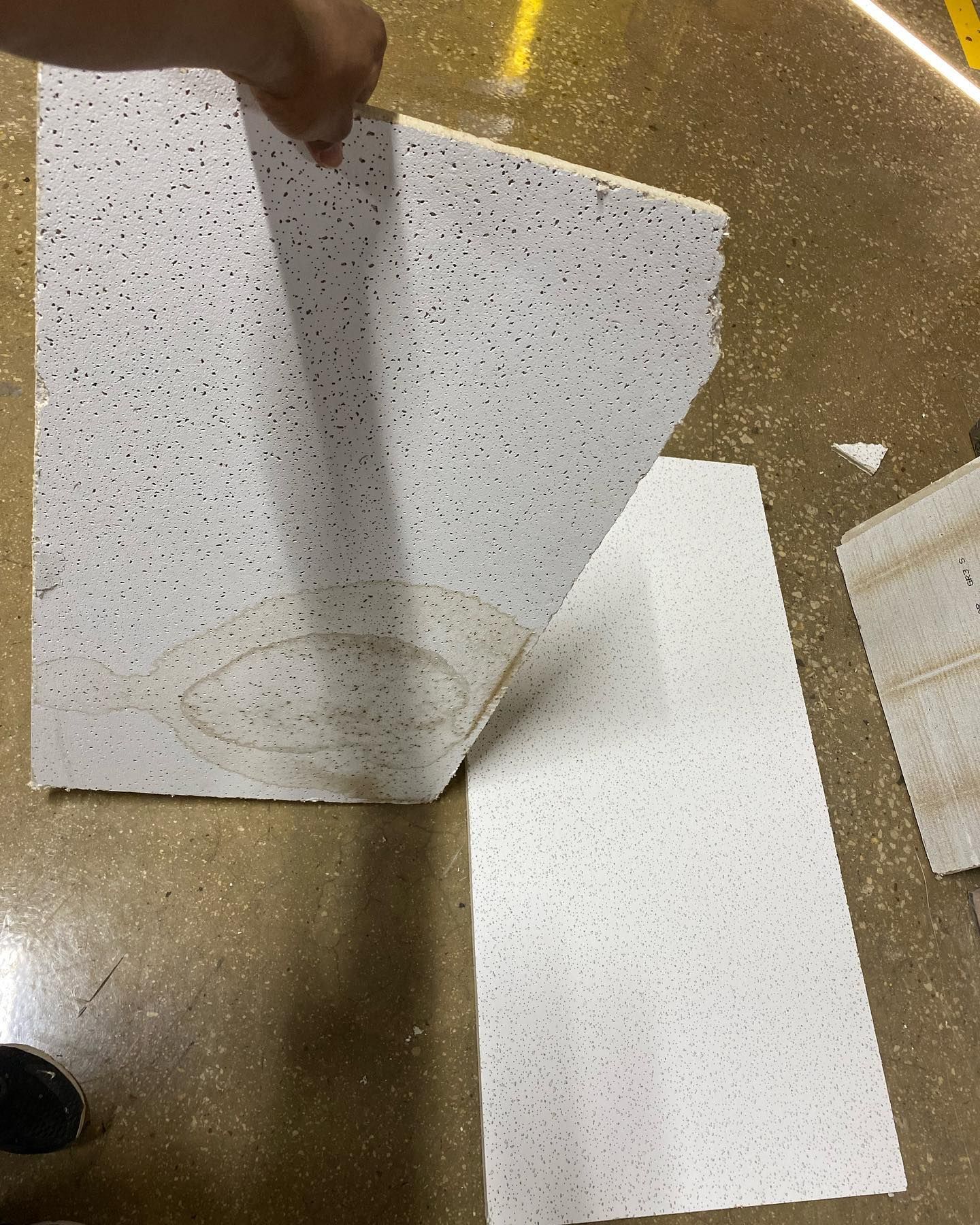 Hand lifting a ceiling tile showing its interior, next to two similar tiles on a speckled floor.