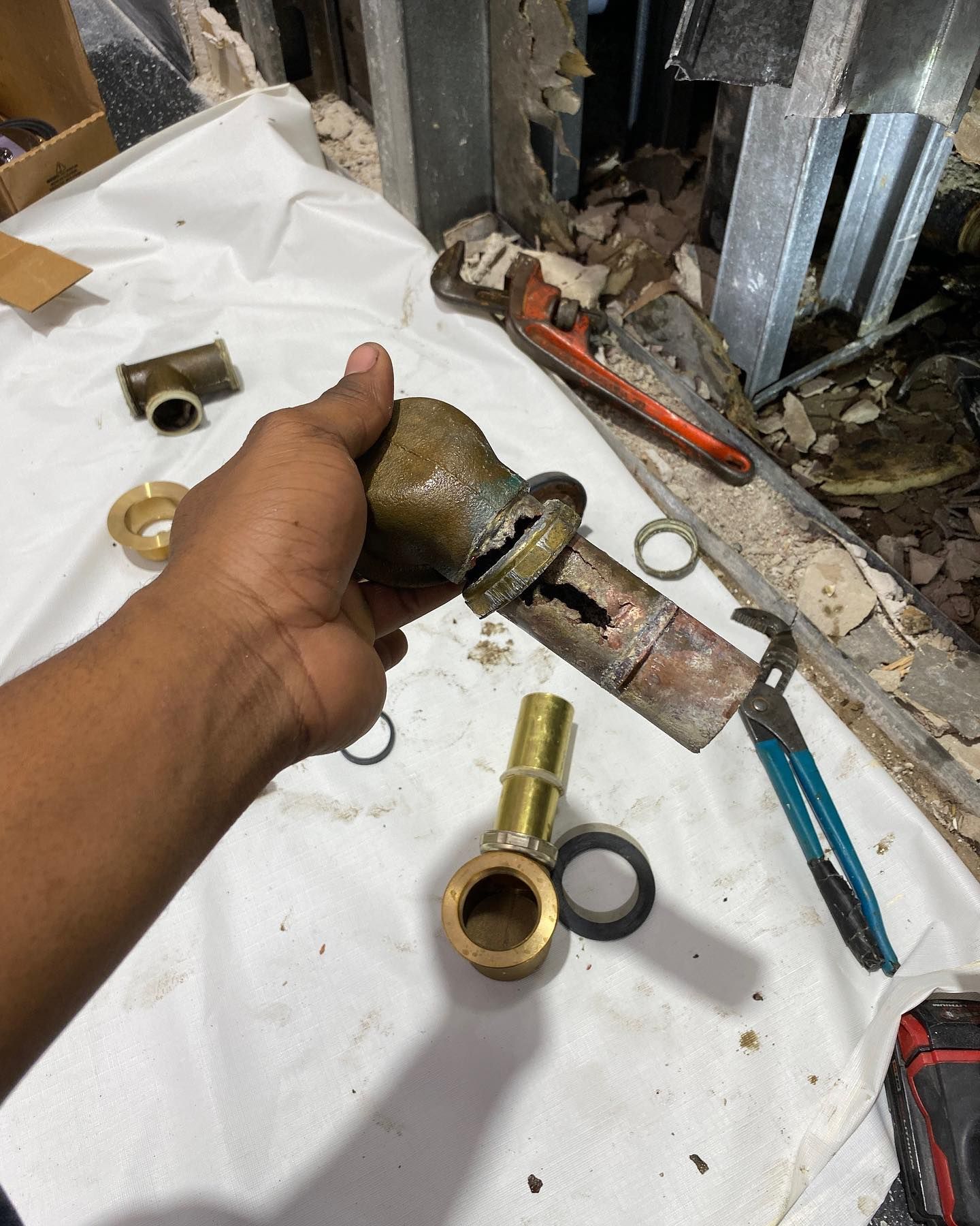 Hand holding a damaged brass pipe with tools, fittings, and debris in a work area.