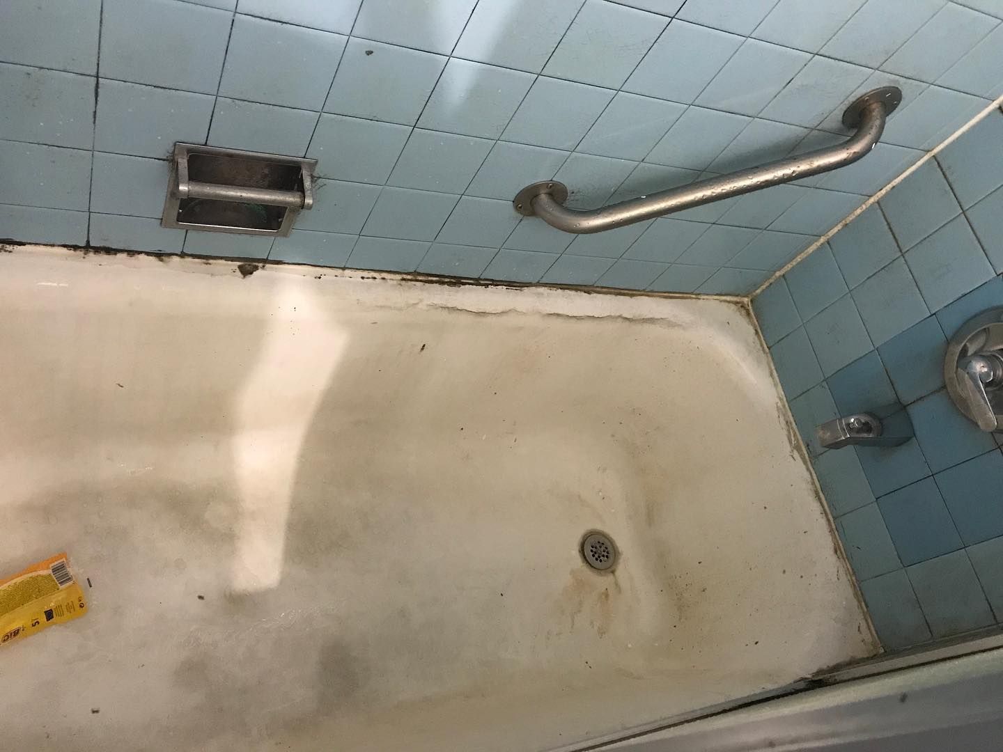 Dirty, stained bathtub with blue tile walls and a grab bar in a bathroom.