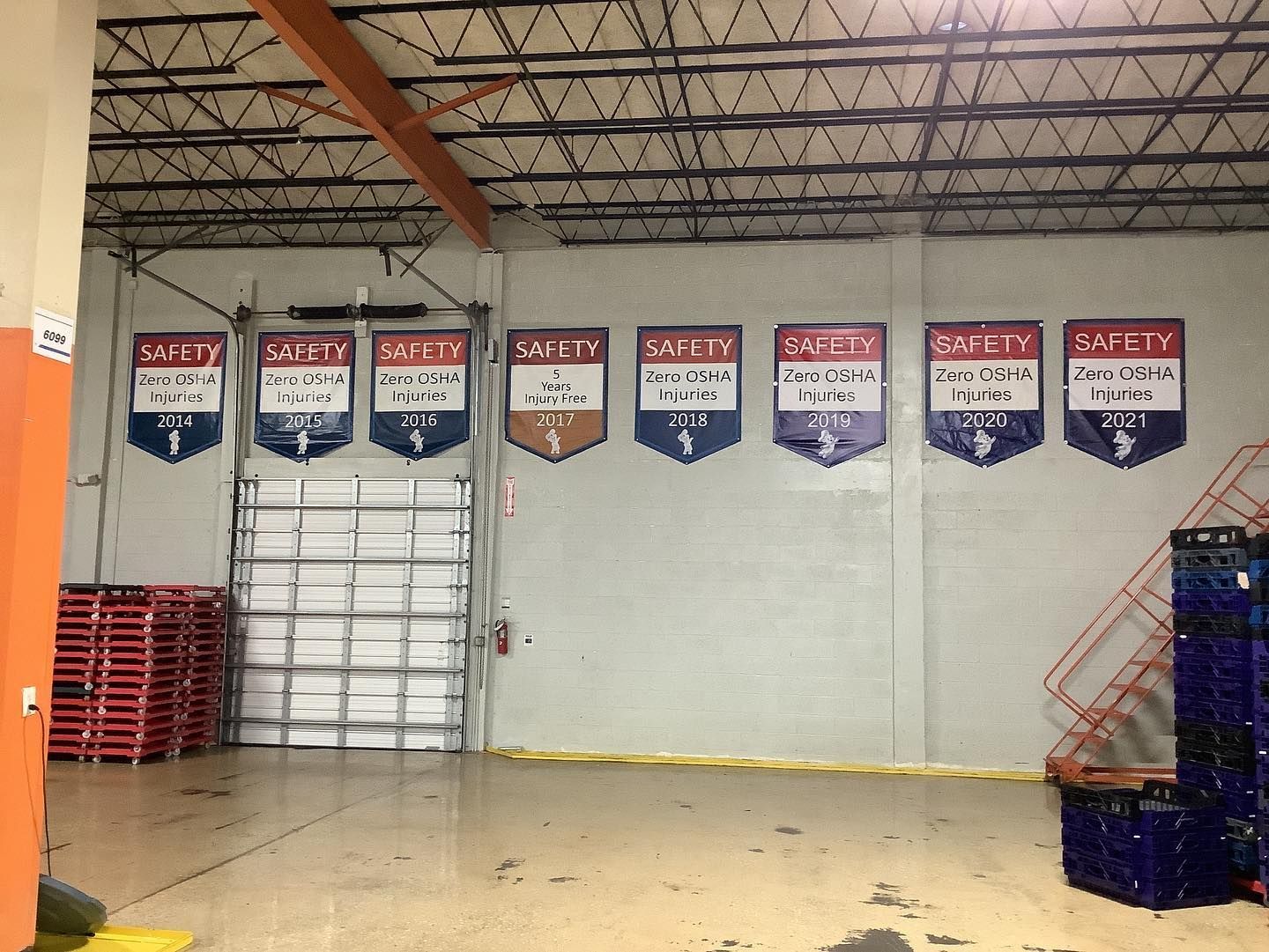 Warehouse interior with safety banners above a closed loading dock door. Red and blue storage containers visible.