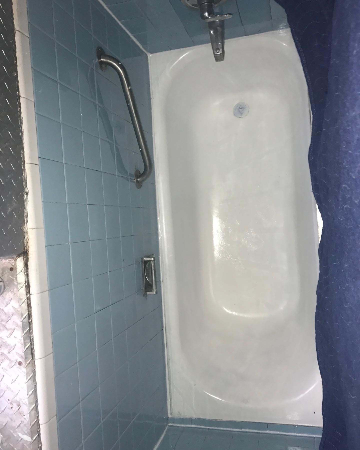 A light-blue tiled bathroom with a white bathtub and a handrail for accessibility.