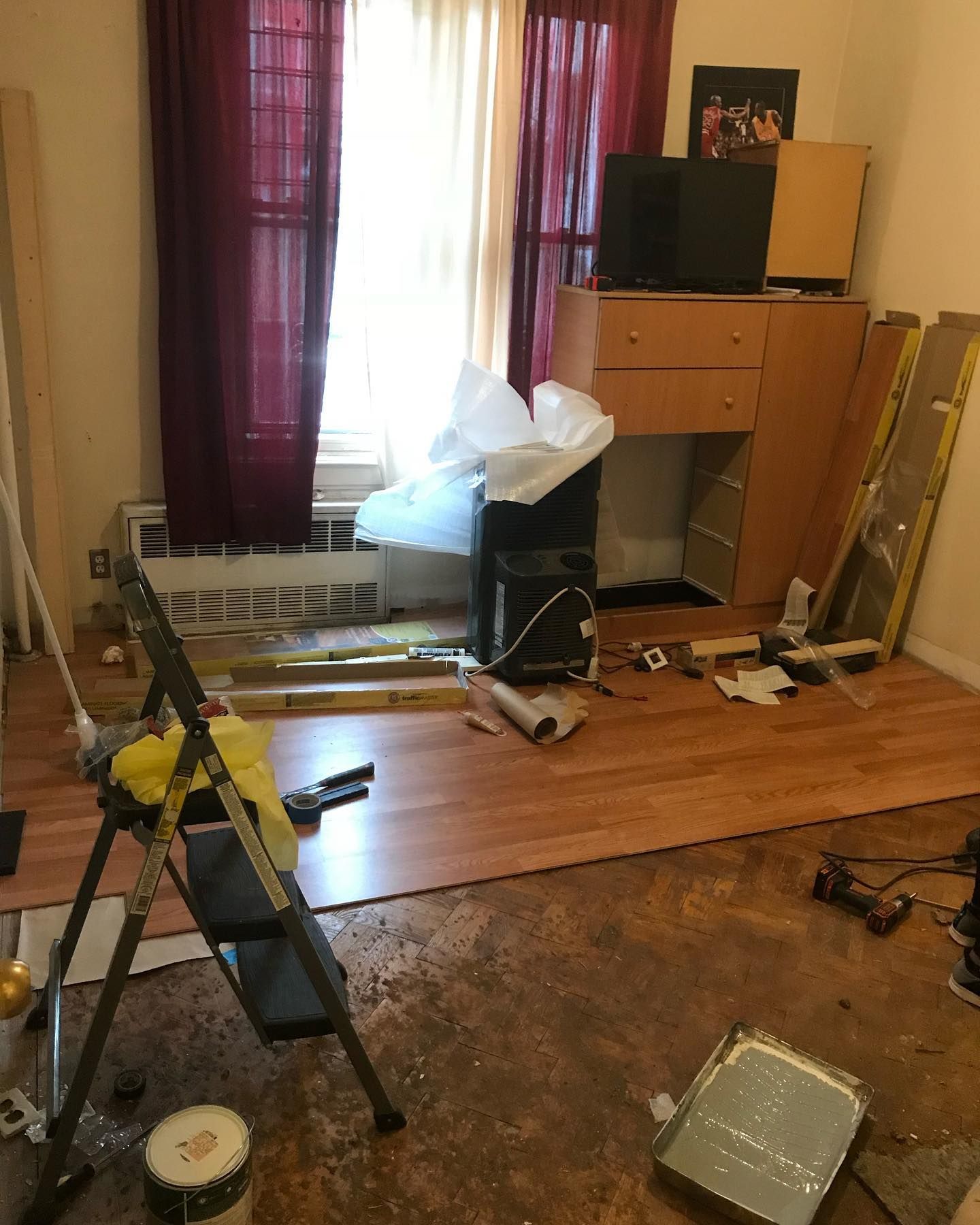 Room with window, burgundy curtains, wood floor installation in progress; tools, ladder, furniture.