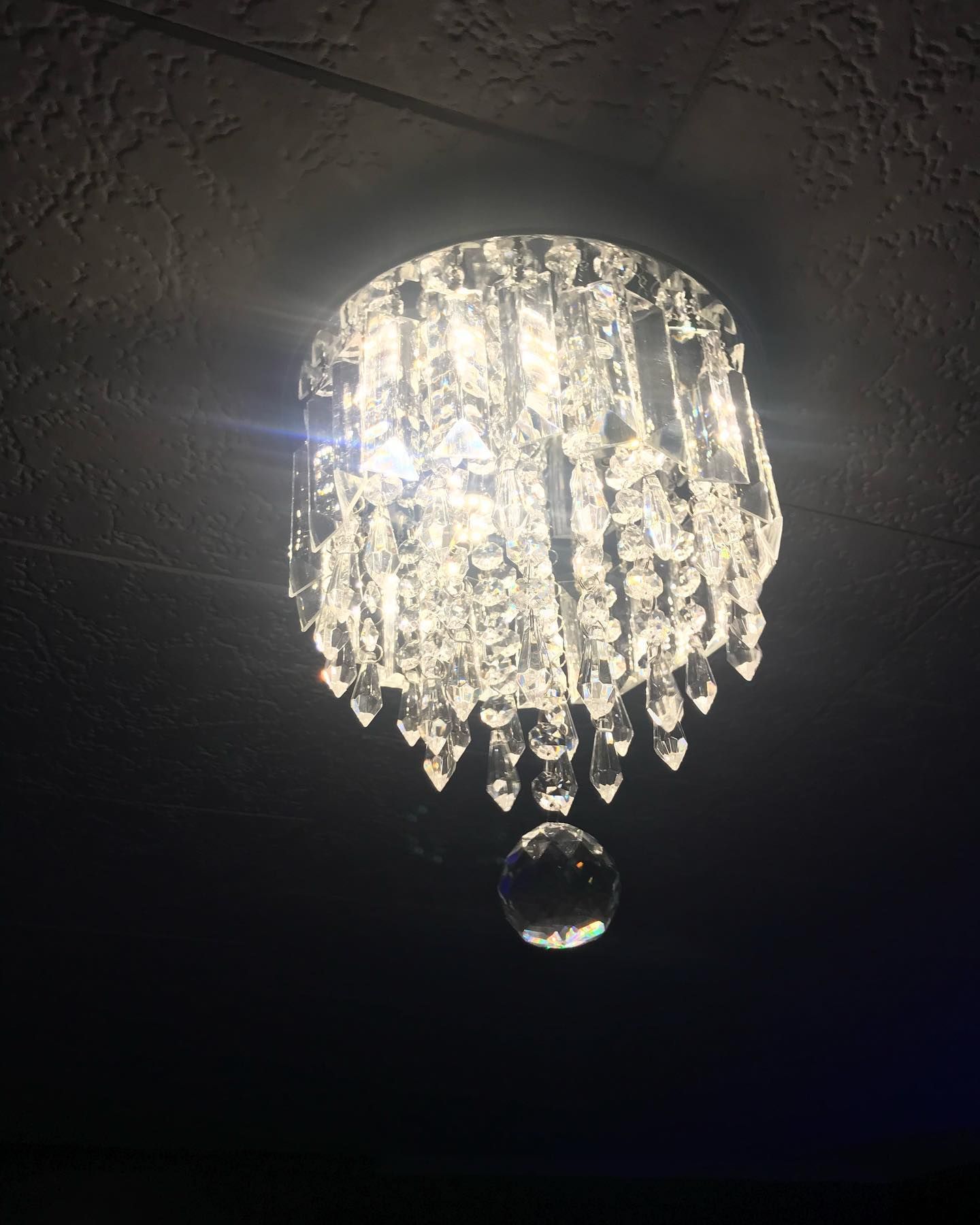 A lit crystal chandelier hangs from a dark ceiling, sparkling brightly.