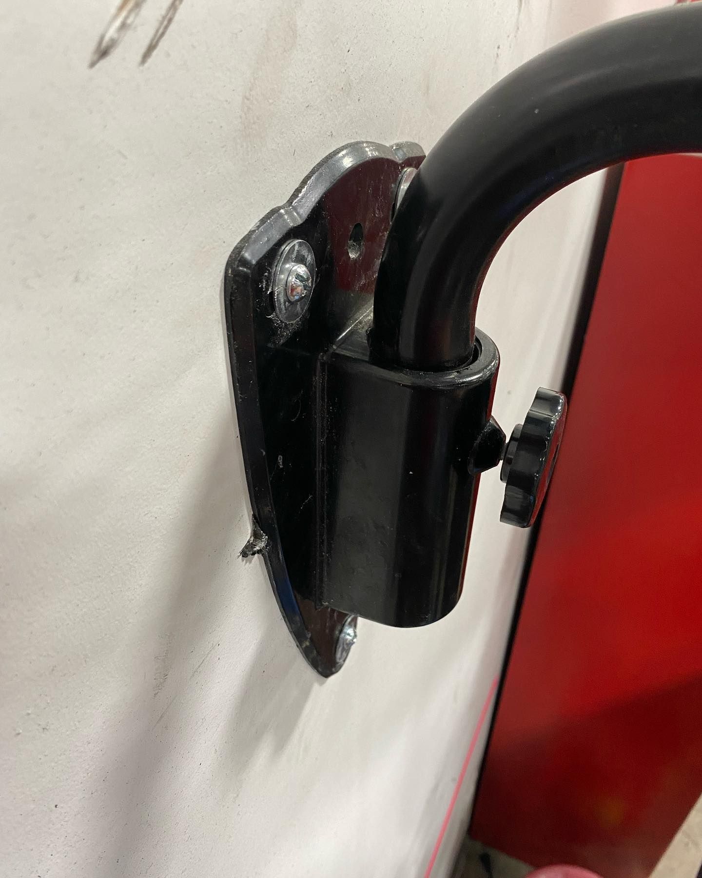 Black bracket with a curved tube attached to a white surface, secured by a screw with a star-shaped handle.