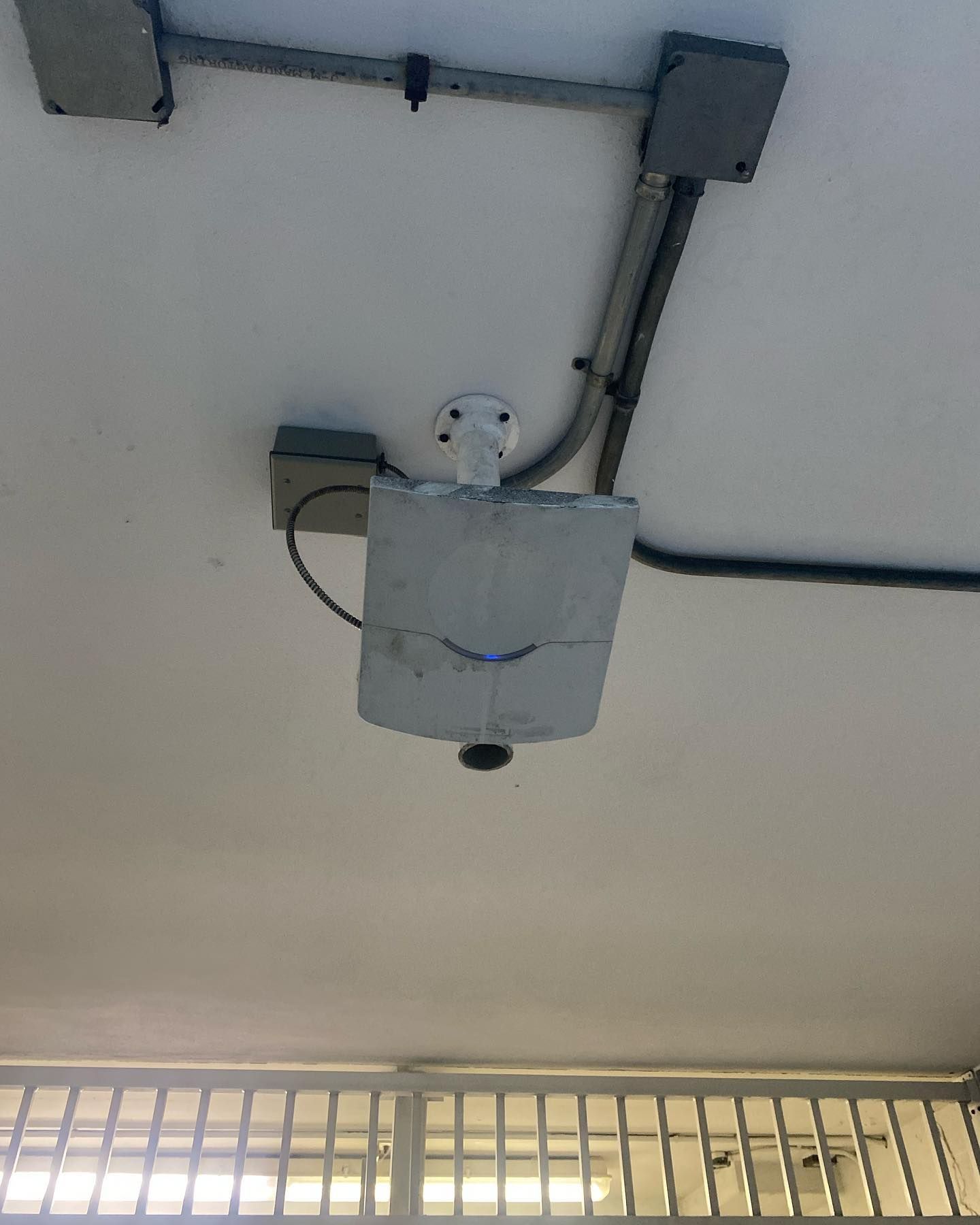 White ceiling-mounted cylinder, possibly a security camera, with conduits and electrical boxes above a metal grate.