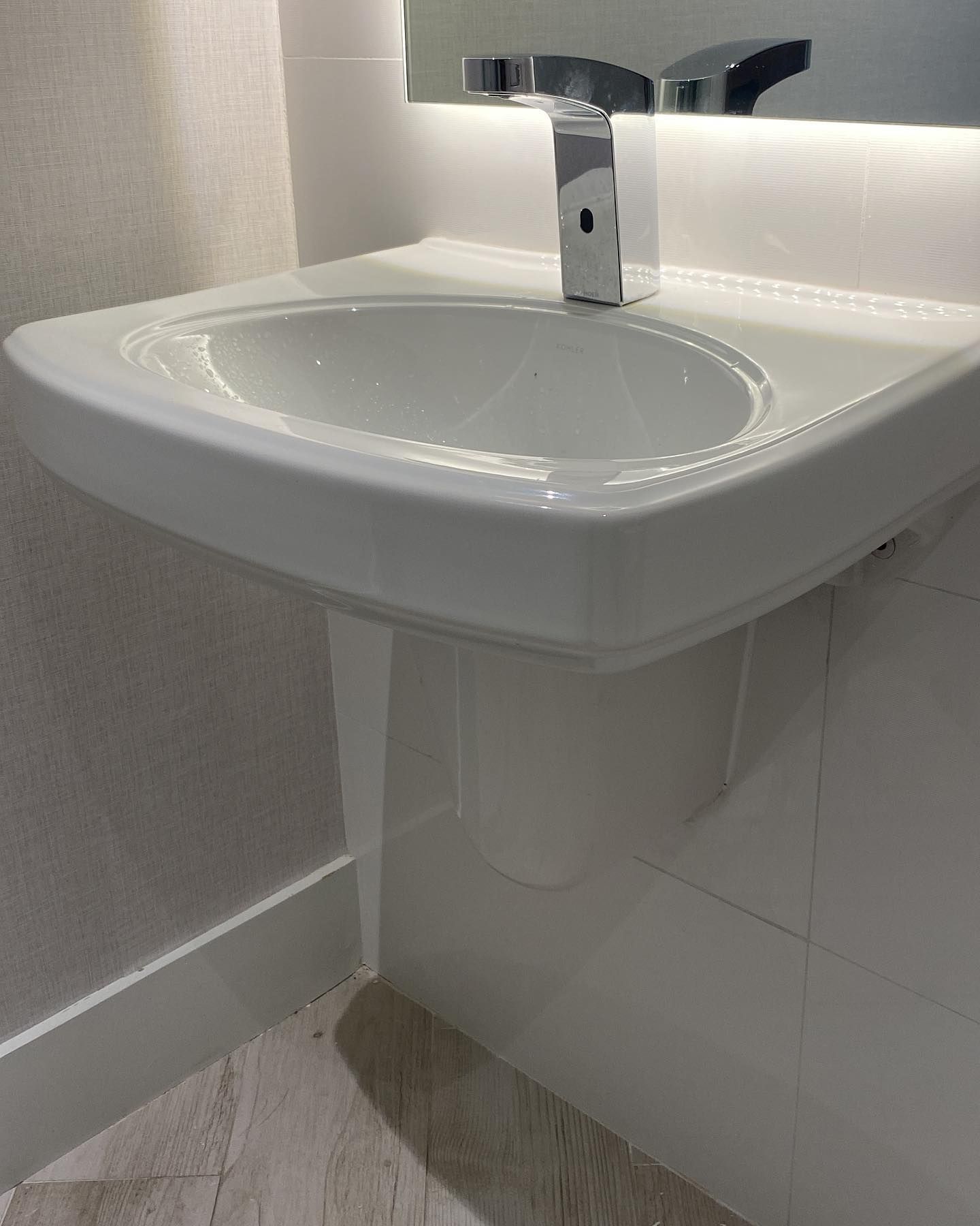 White wall-mounted bathroom sink with chrome faucet, installed over white tiles and a light-colored floor.
