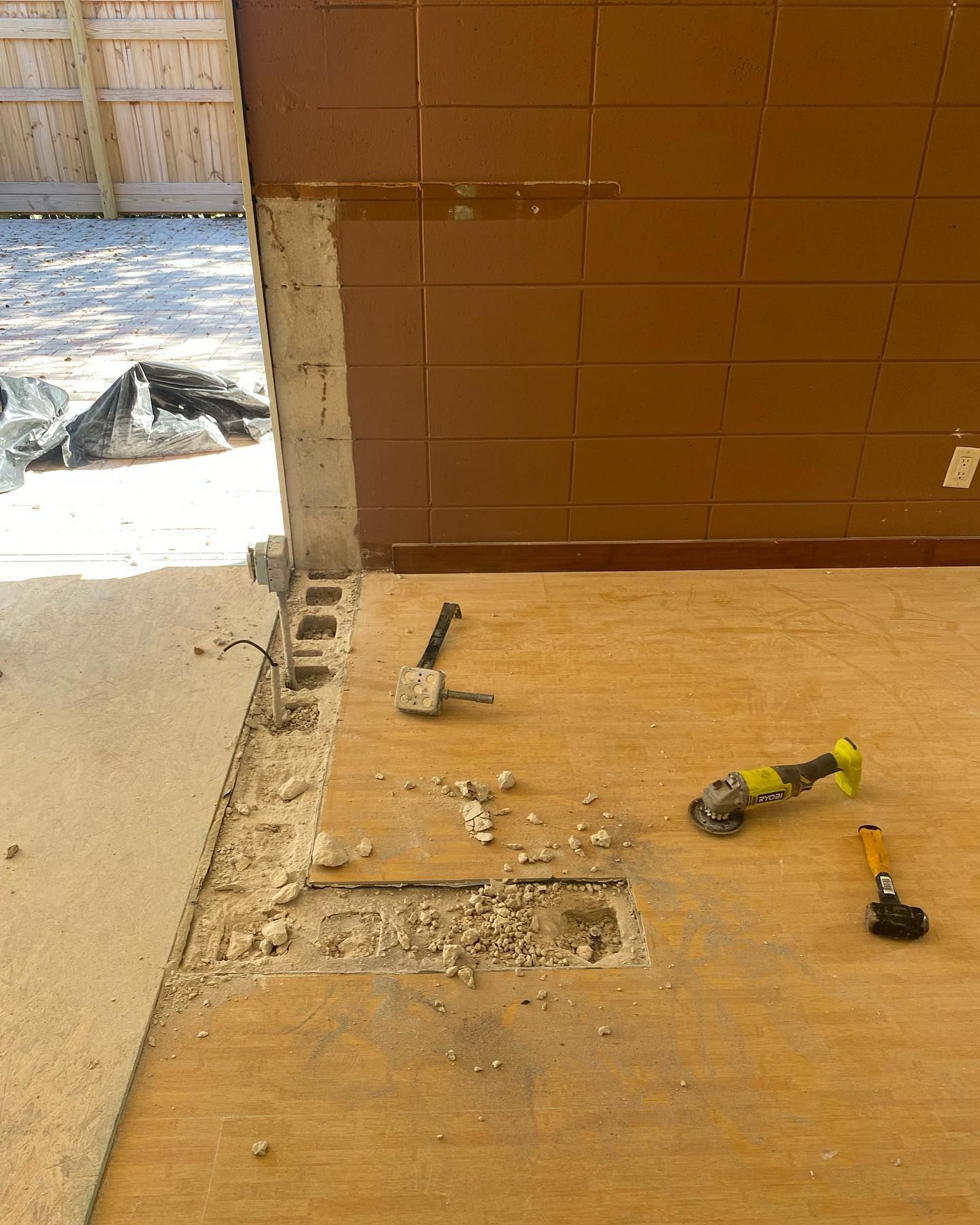 Construction site. A wall being partially demolished. Tools lay on plywood flooring. Beige, brown, and gray. Outdoors.