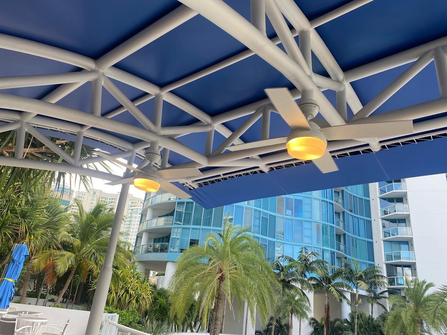 Blue canopy with fans over pool area, palm trees, and a high-rise building.