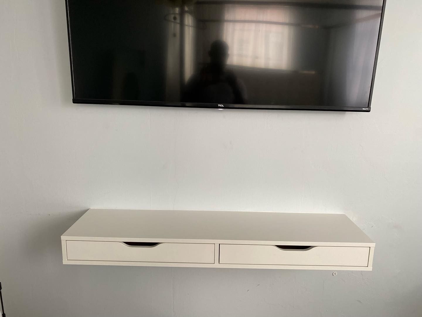 A white floating drawer unit with two drawers under a wall-mounted black TV on a white wall.