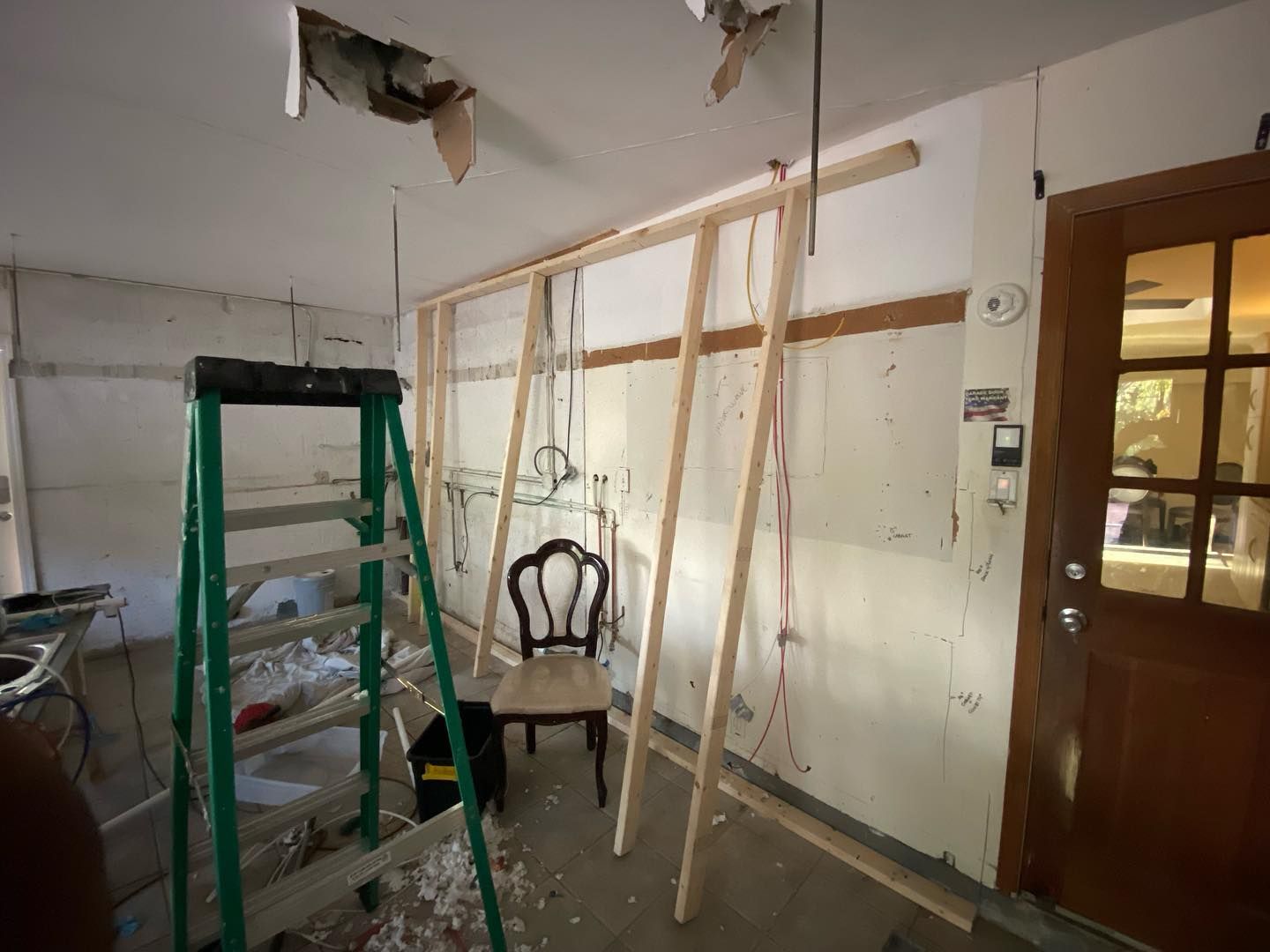 Construction in a room with a ladder, wood framing, a chair, and a doorway. White walls, exposed wires.