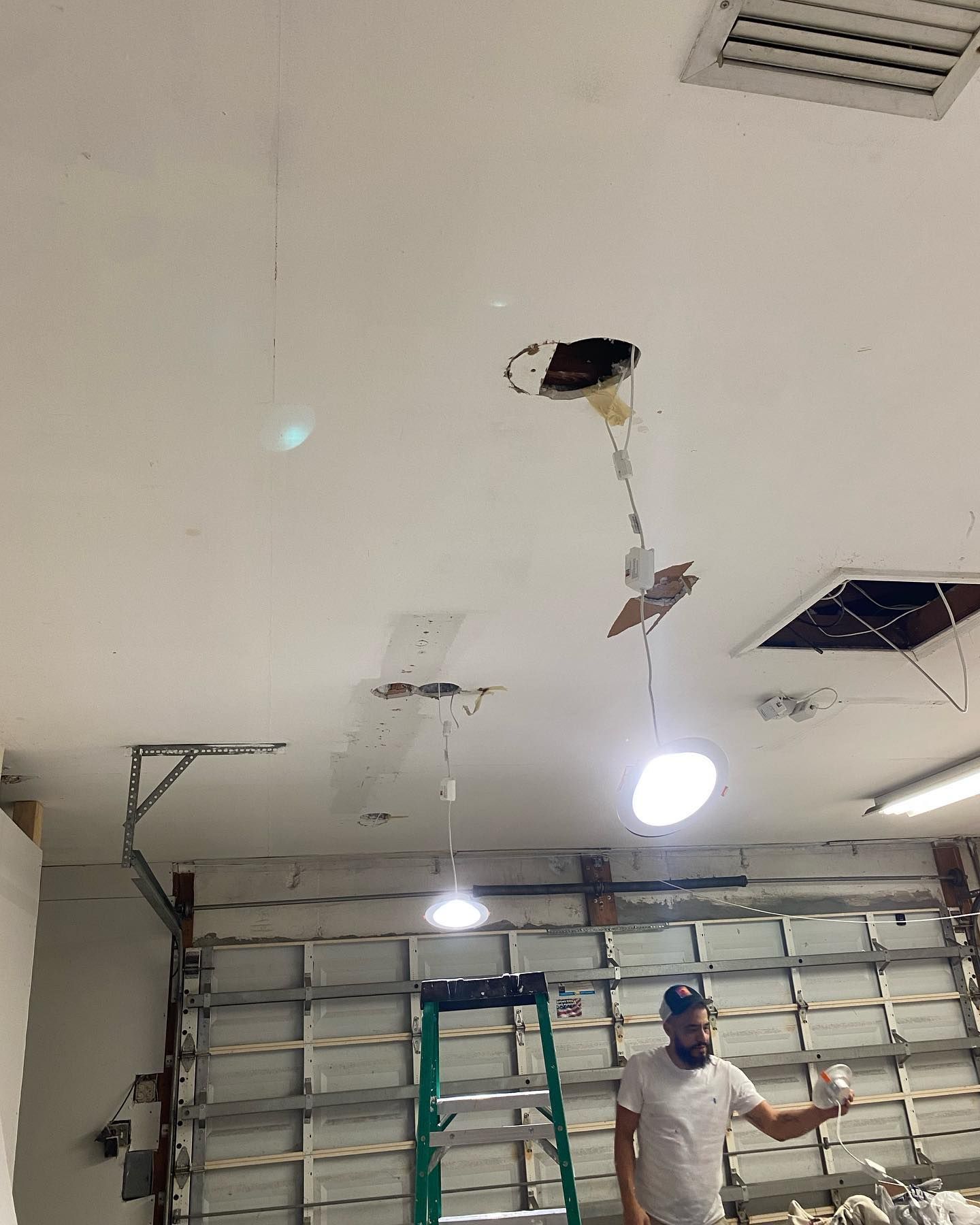Man in garage, working on ceiling light fixture with ladder nearby, other light fixtures visible.