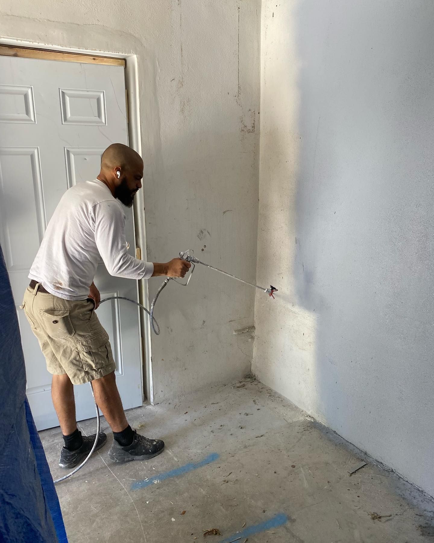 Man in shorts spray-painting a wall in a room with a door, light blue color, concrete floor.