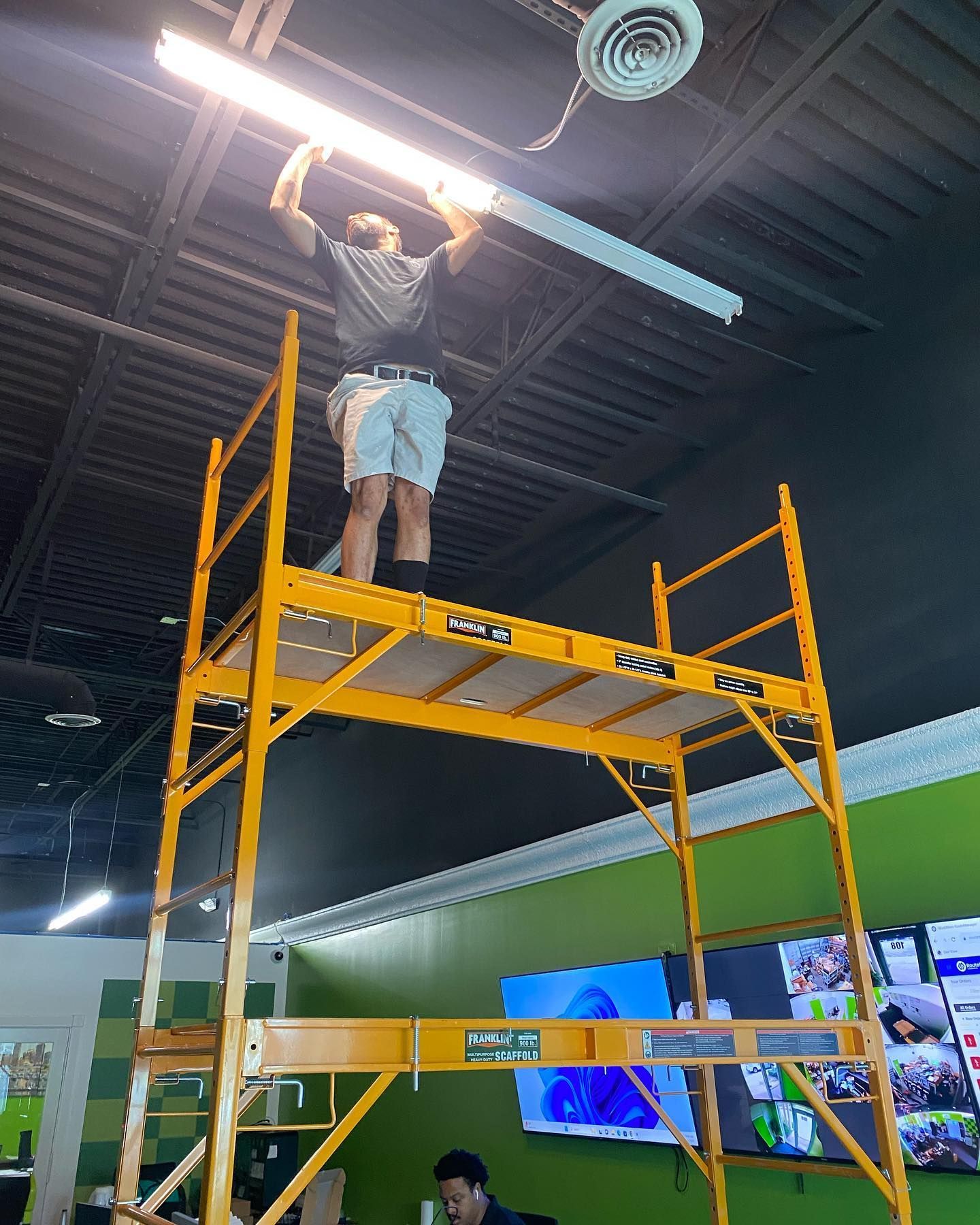 Man on a yellow scaffold installing a light fixture in a room with green and black walls.