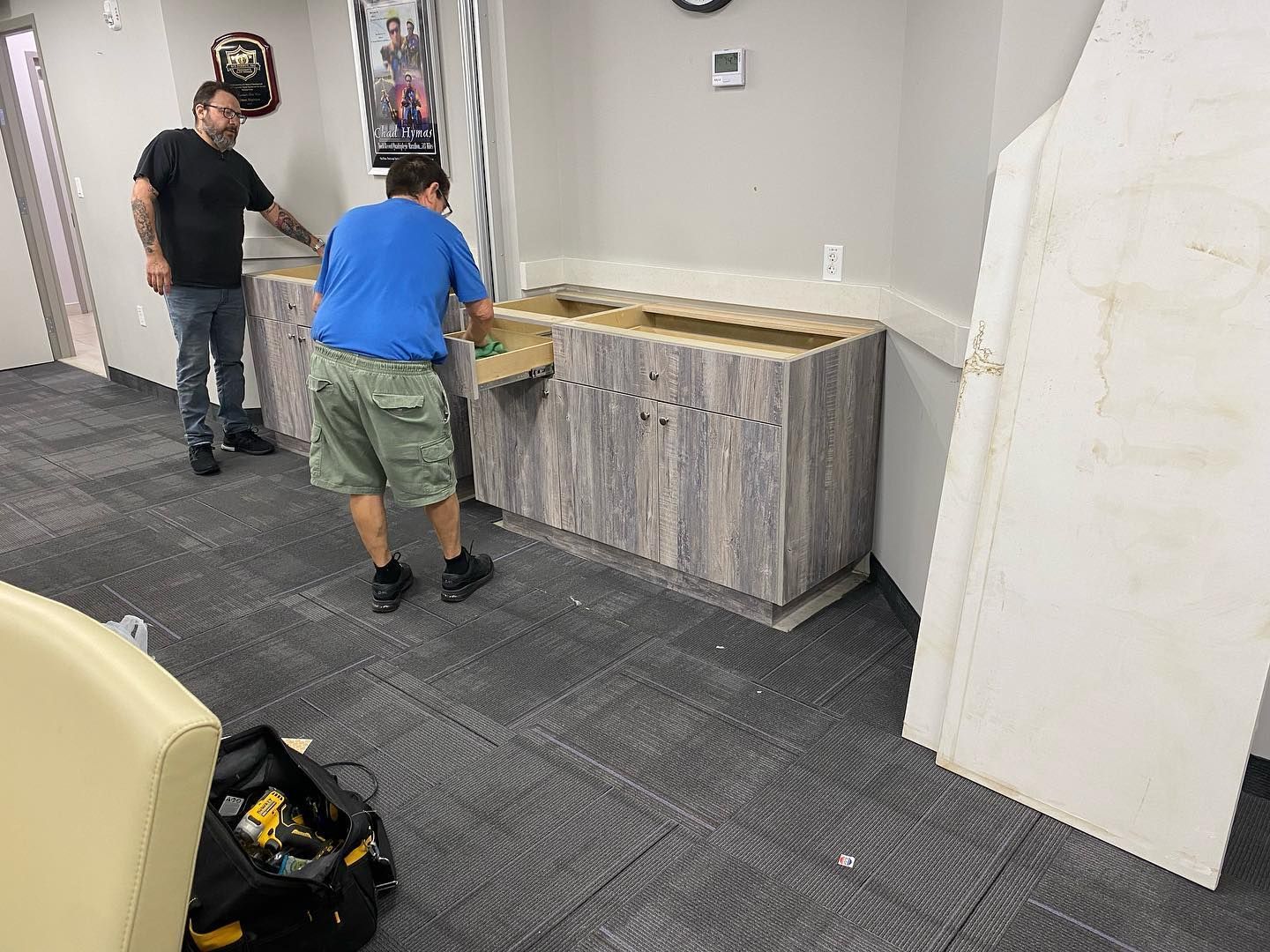 Two men installing a cabinet in an office. One man is holding a box. Grey and white color scheme.
