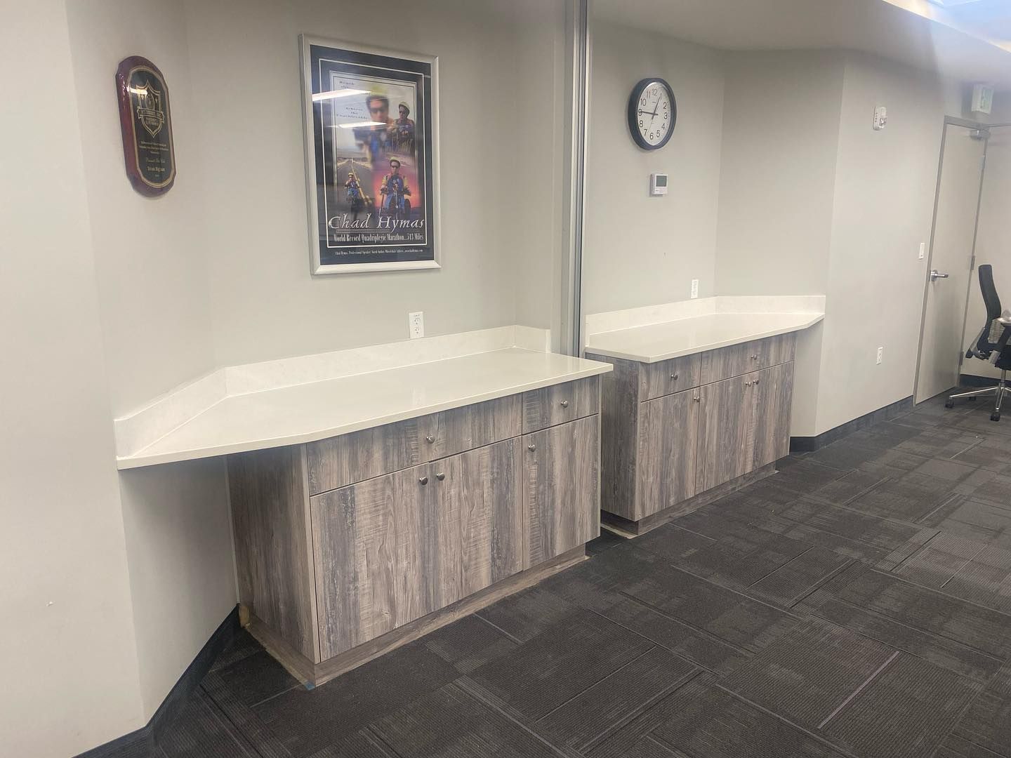 Two gray-toned wooden cabinets with white countertops in an office hallway. Movie poster and clock on the wall.