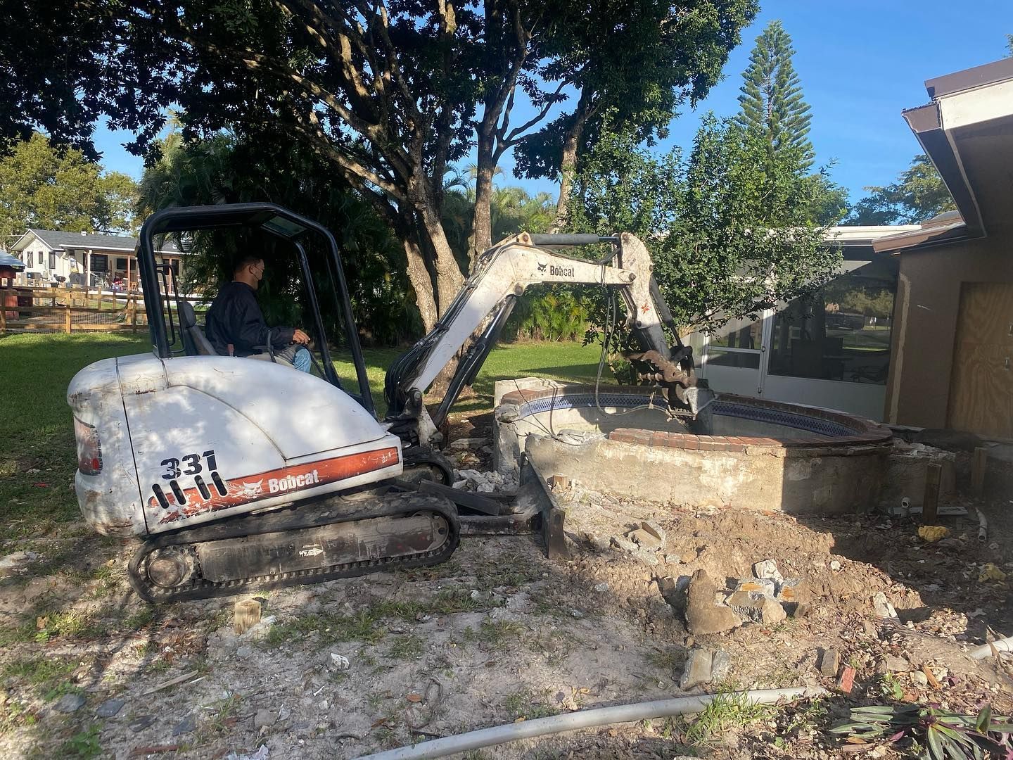 A person operates a Bobcat excavator, demolishing a concrete structure near a building in a yard.