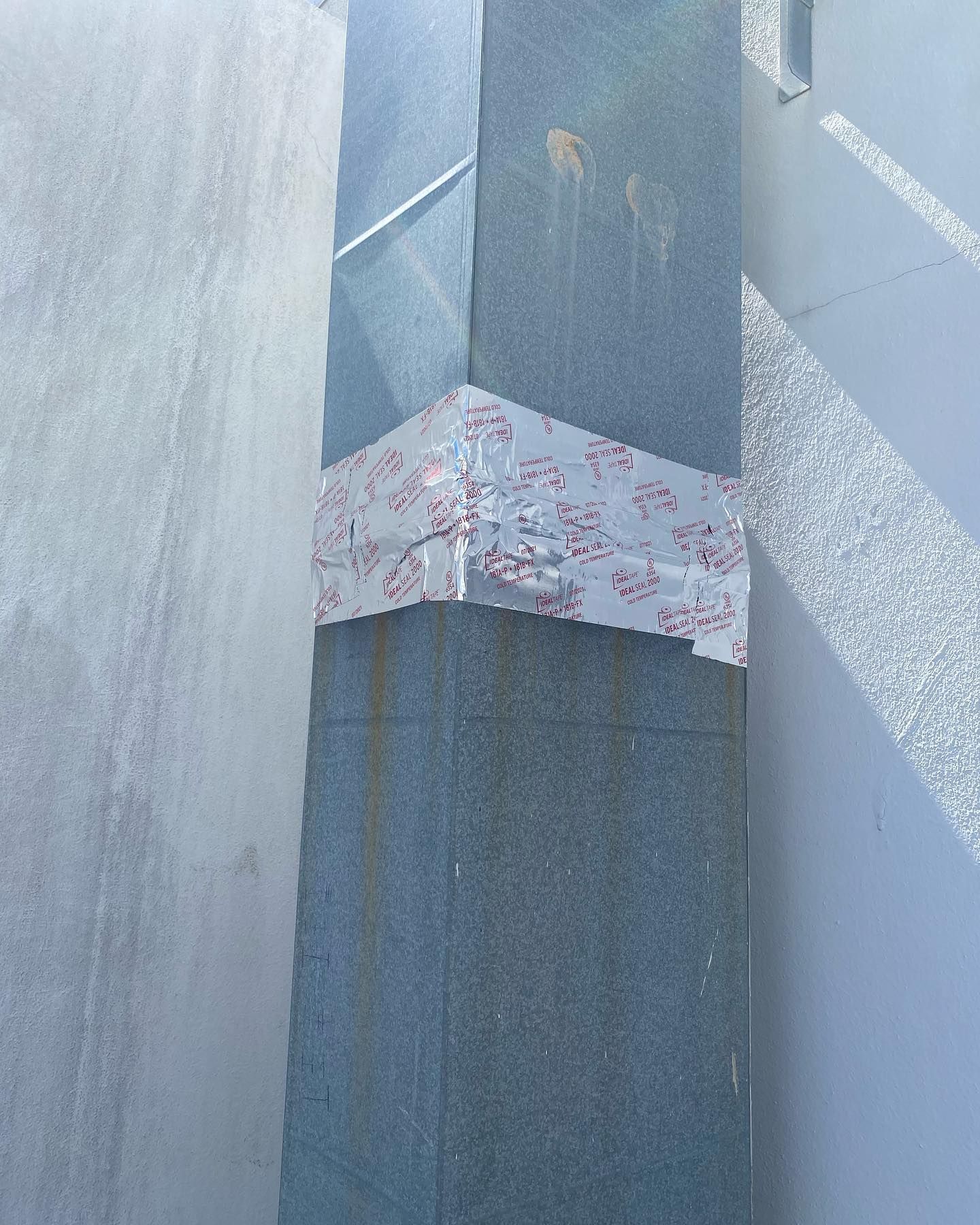 Close-up of a silver metal duct sealed with reflective tape, near a wall and a flat surface.