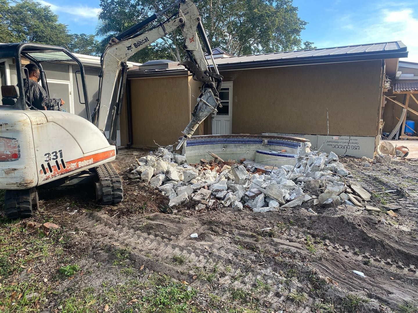 A mini excavator demolishes a concrete structure in front of a beige building on a sunny day.