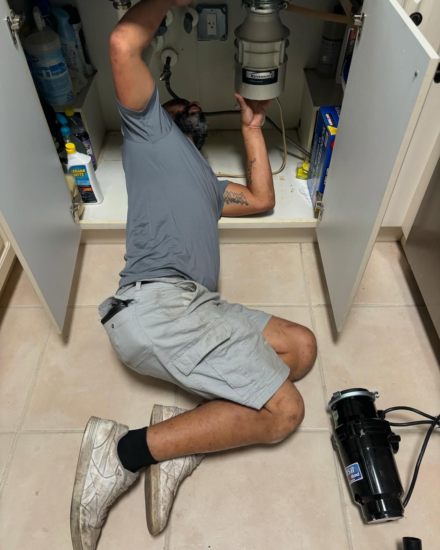 Man kneeling under kitchen sink, working on garbage disposal. Beige and gray tones.