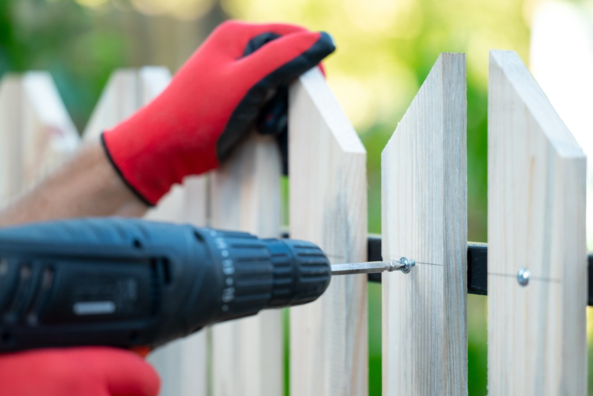 Person in red gloves using a drill to attach a wooden fence picket to a horizontal rail outdoors.