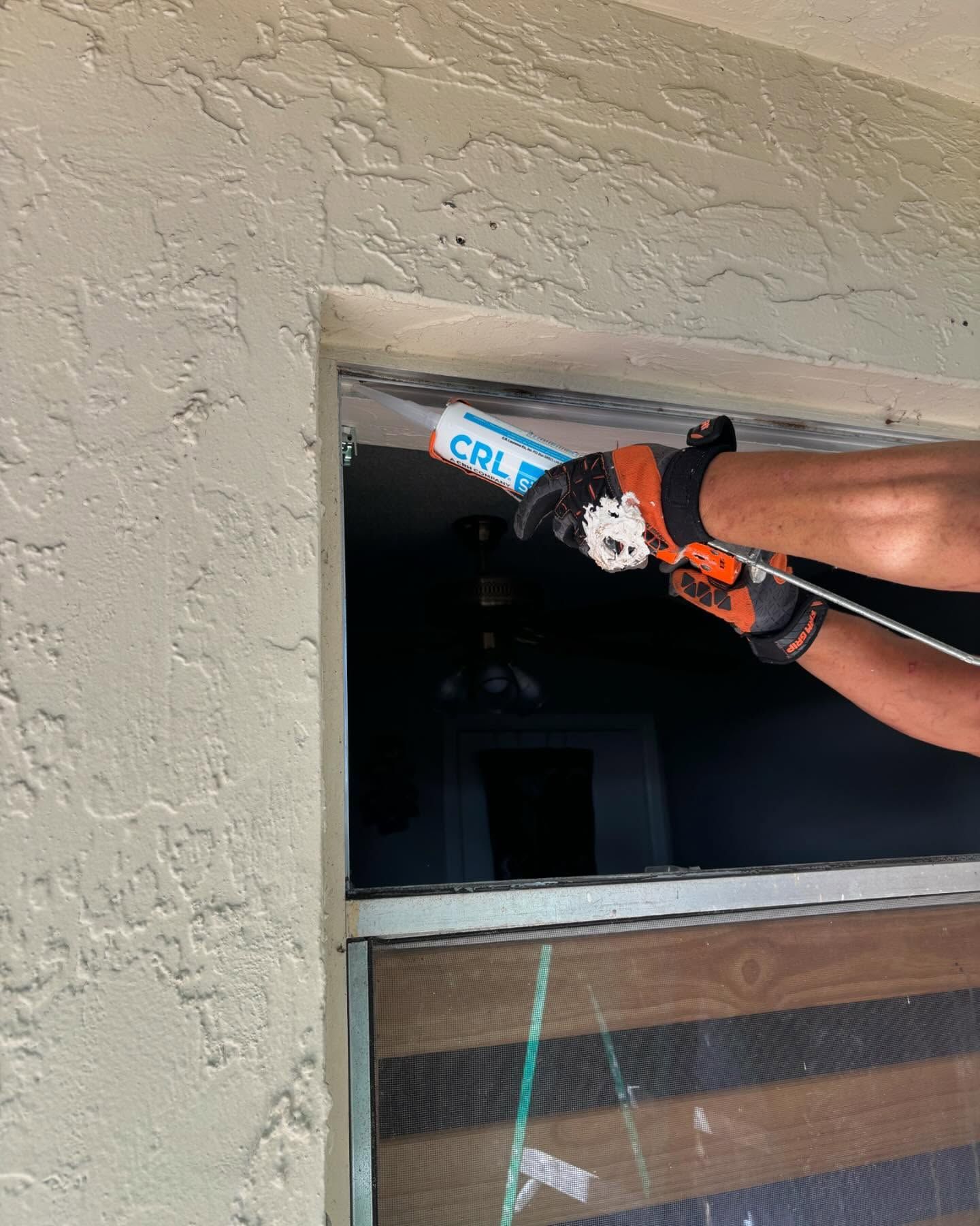 Person using a caulk gun to seal around a window frame, outdoors. Wearing work gloves.