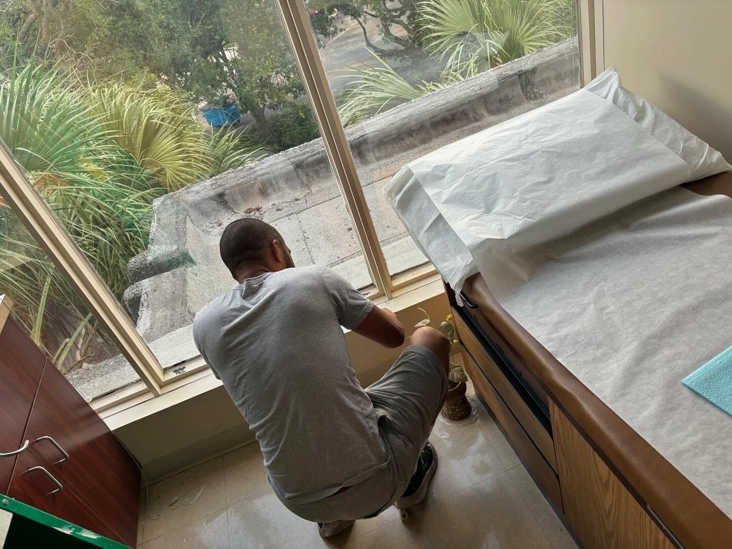 Man cleaning a window in a medical office; beside an examination bed with white linens, outside greenery visible.
