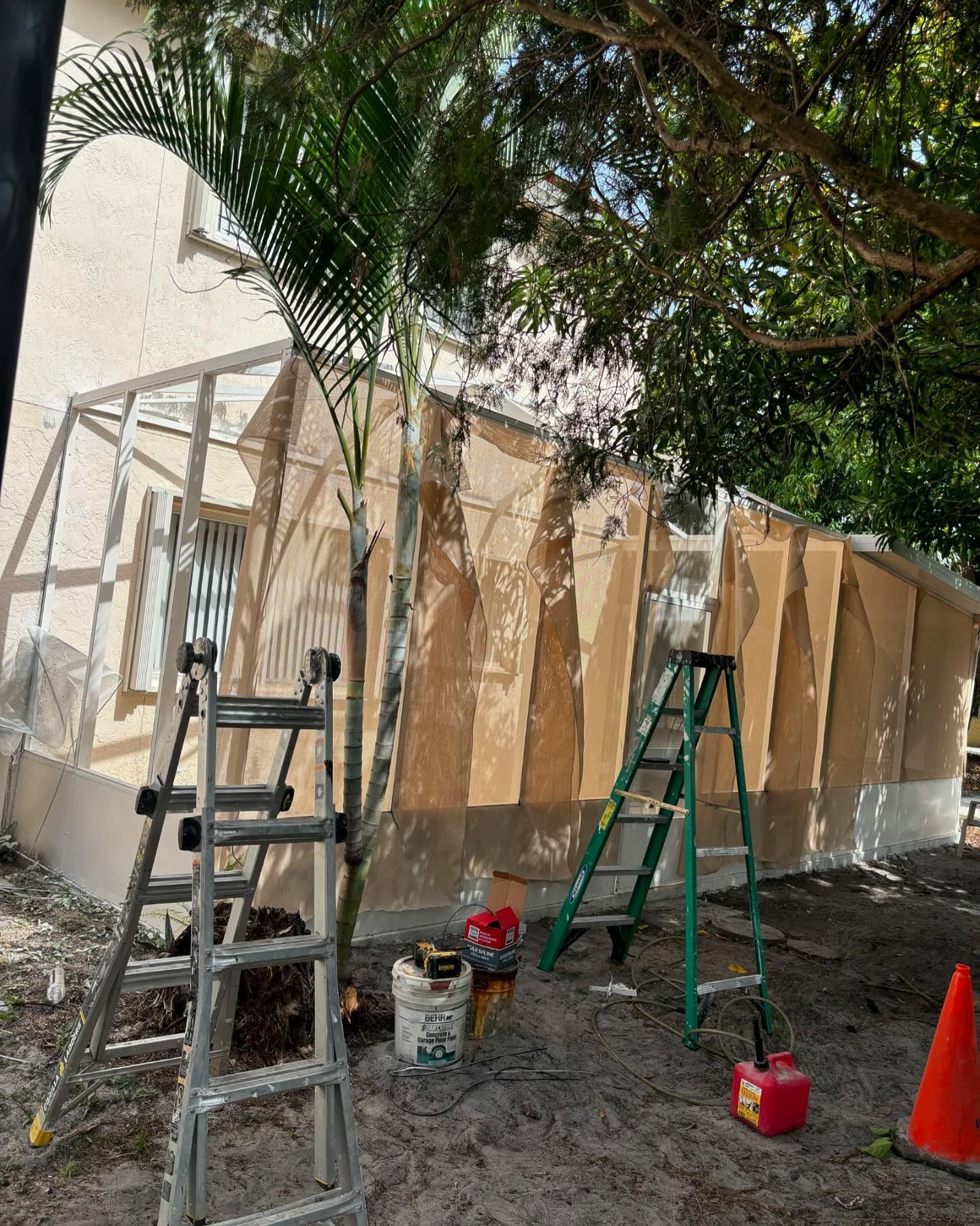 Construction site: a building with partially installed beige corrugated panels. Ladders, tools, and a palm tree are present.