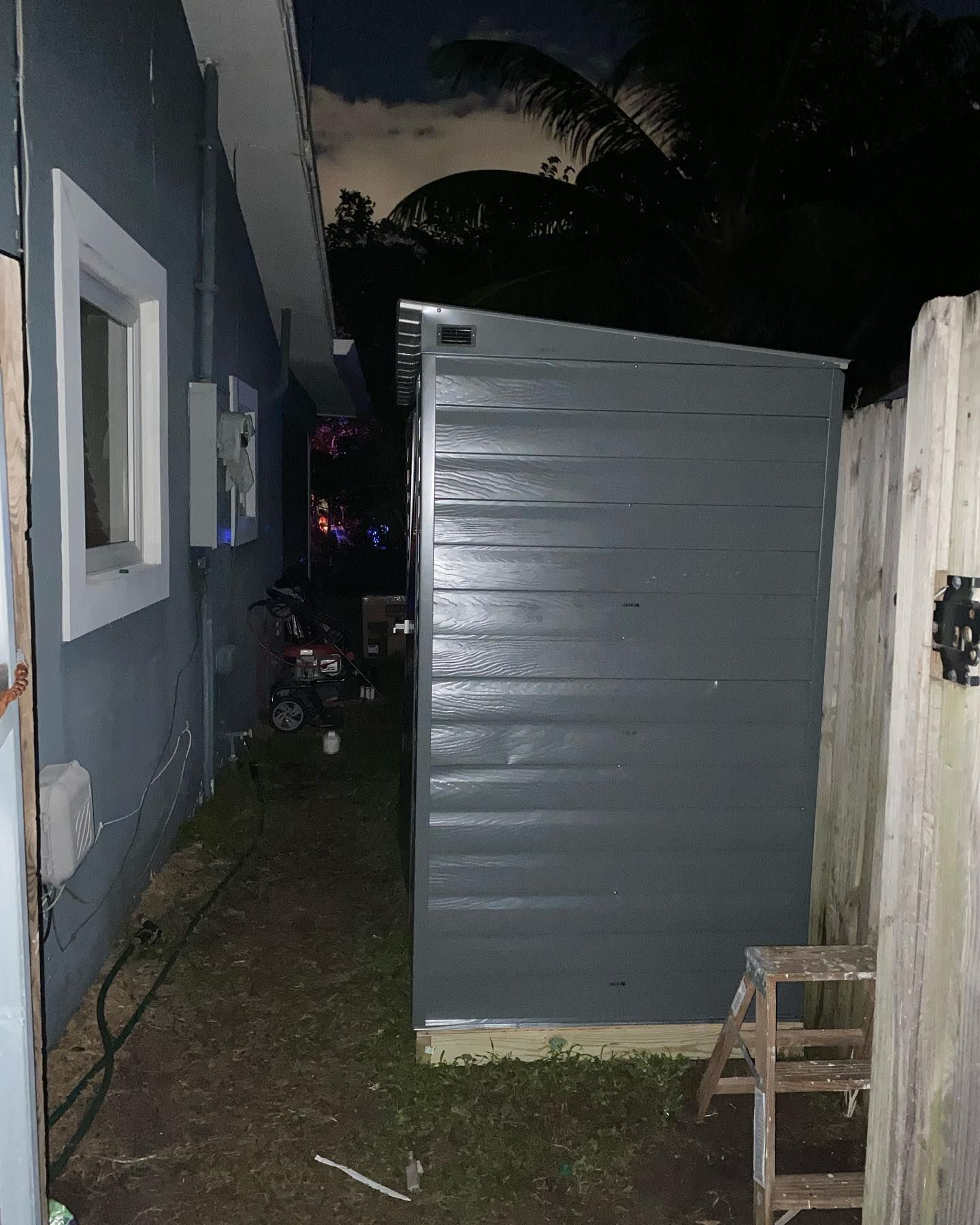 A gray shed next to a blue house, with a small step stool nearby. Dark backyard setting.