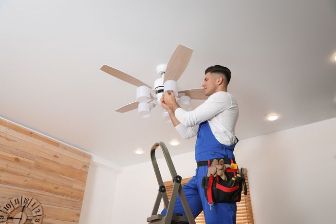 Man in blue overalls on ladder, installing light bulbs on a ceiling fan in a room.