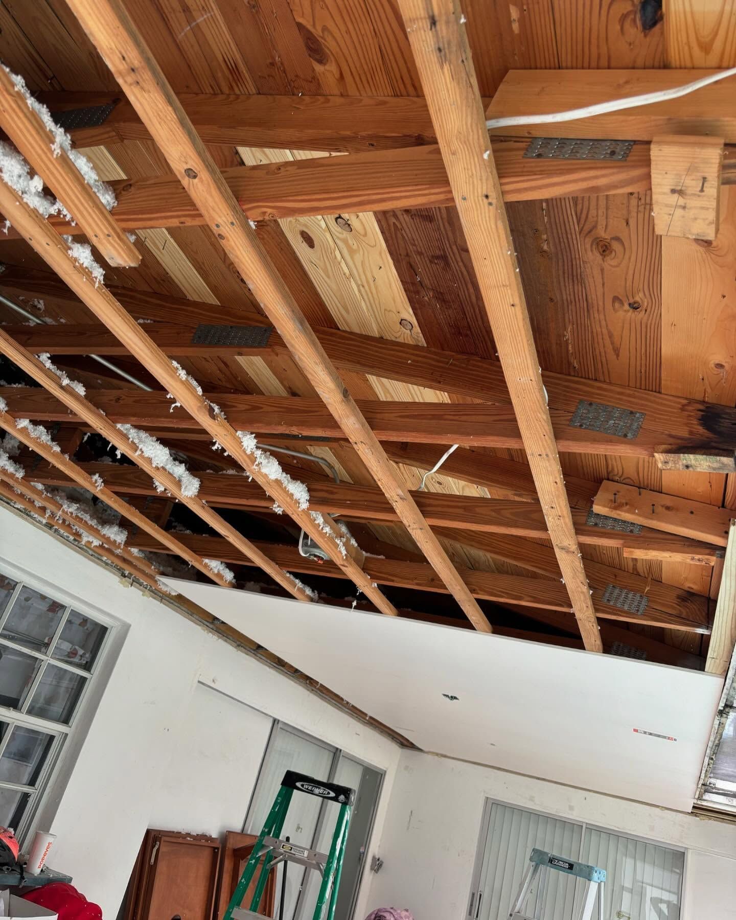 Interior view of ceiling under construction, showing exposed wooden beams, insulation, and drywall being installed.