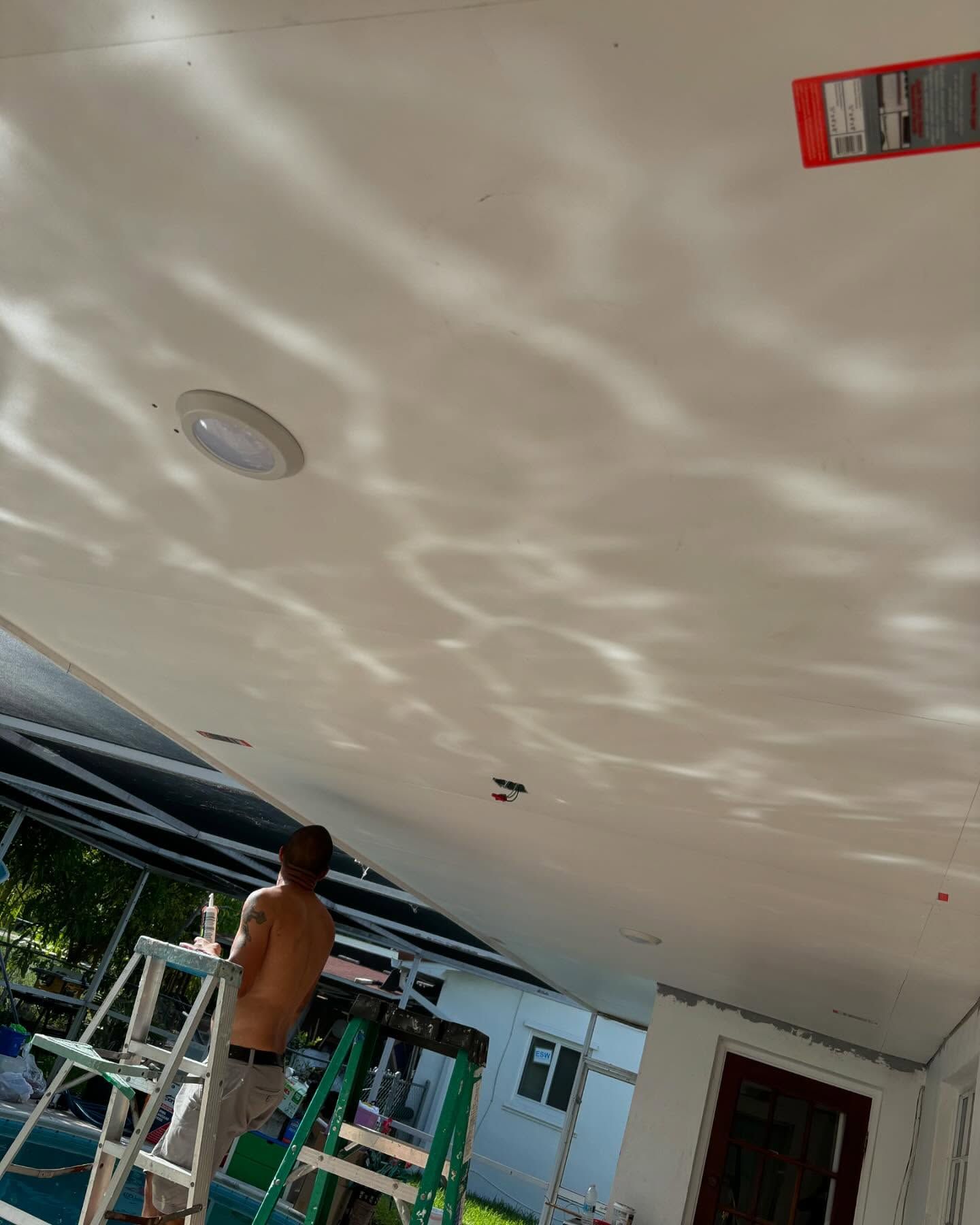 A man painting a white ceiling outdoors, standing on a ladder near a pool.