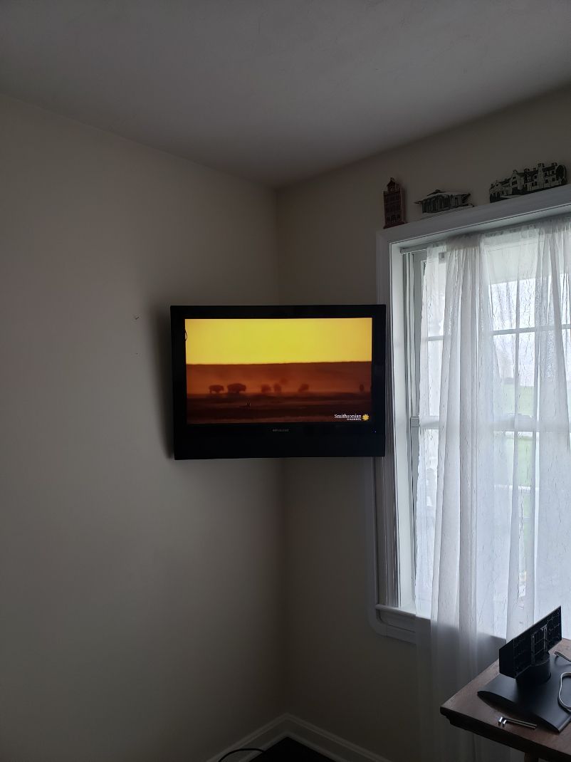 Wall-mounted TV showing a sunset scene beside a window with sheer curtains in a small room