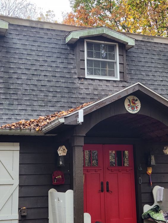 Red front door under a dark shingled porch with a dormer window and autumn leaves on the roof