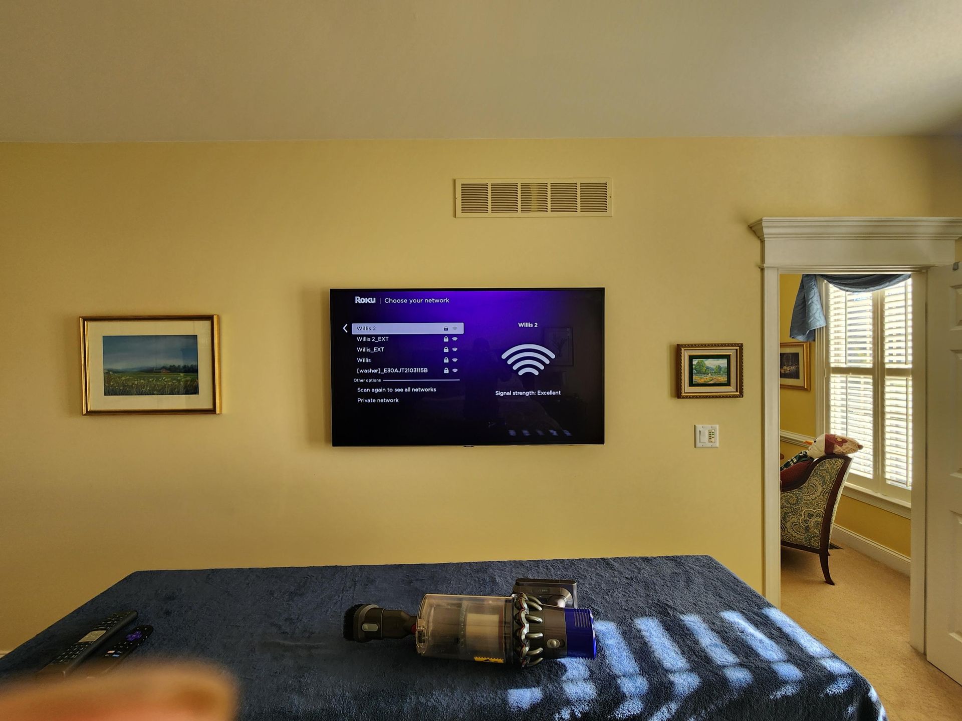 Bedroom with wall-mounted TV, framed art, bed, and open doorway to a bright adjoining room.