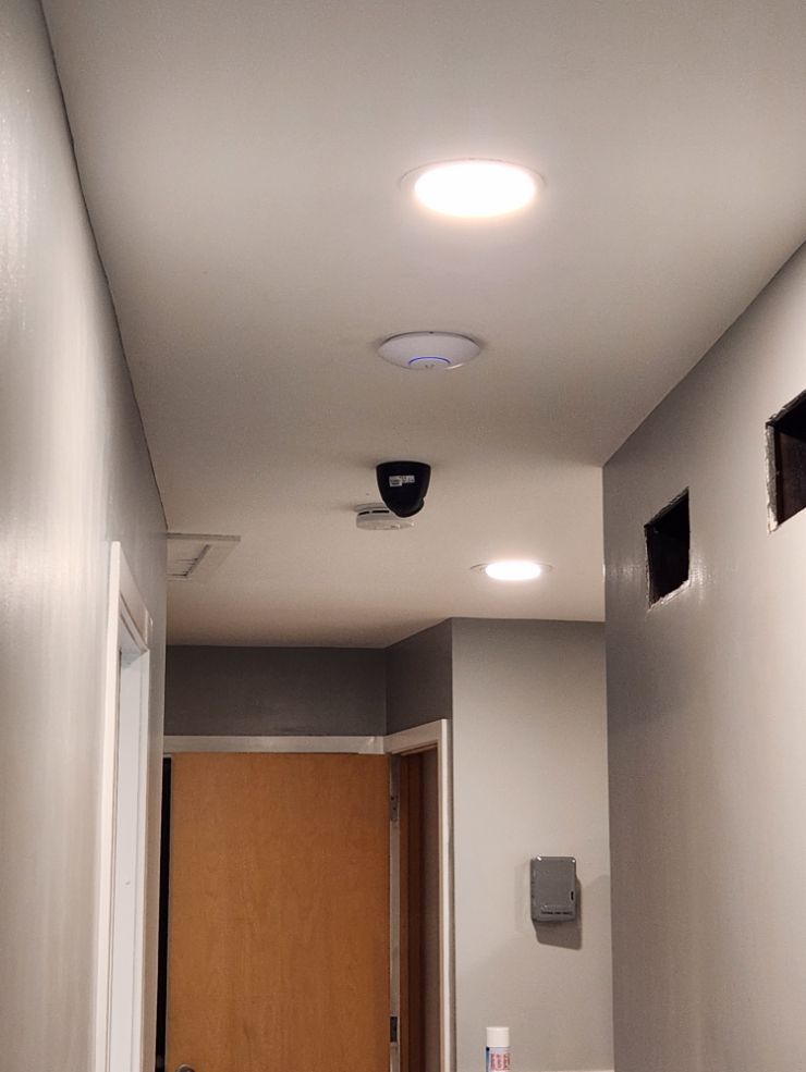 Narrow hallway with ceiling lights, a security camera, and a wooden door at the end.